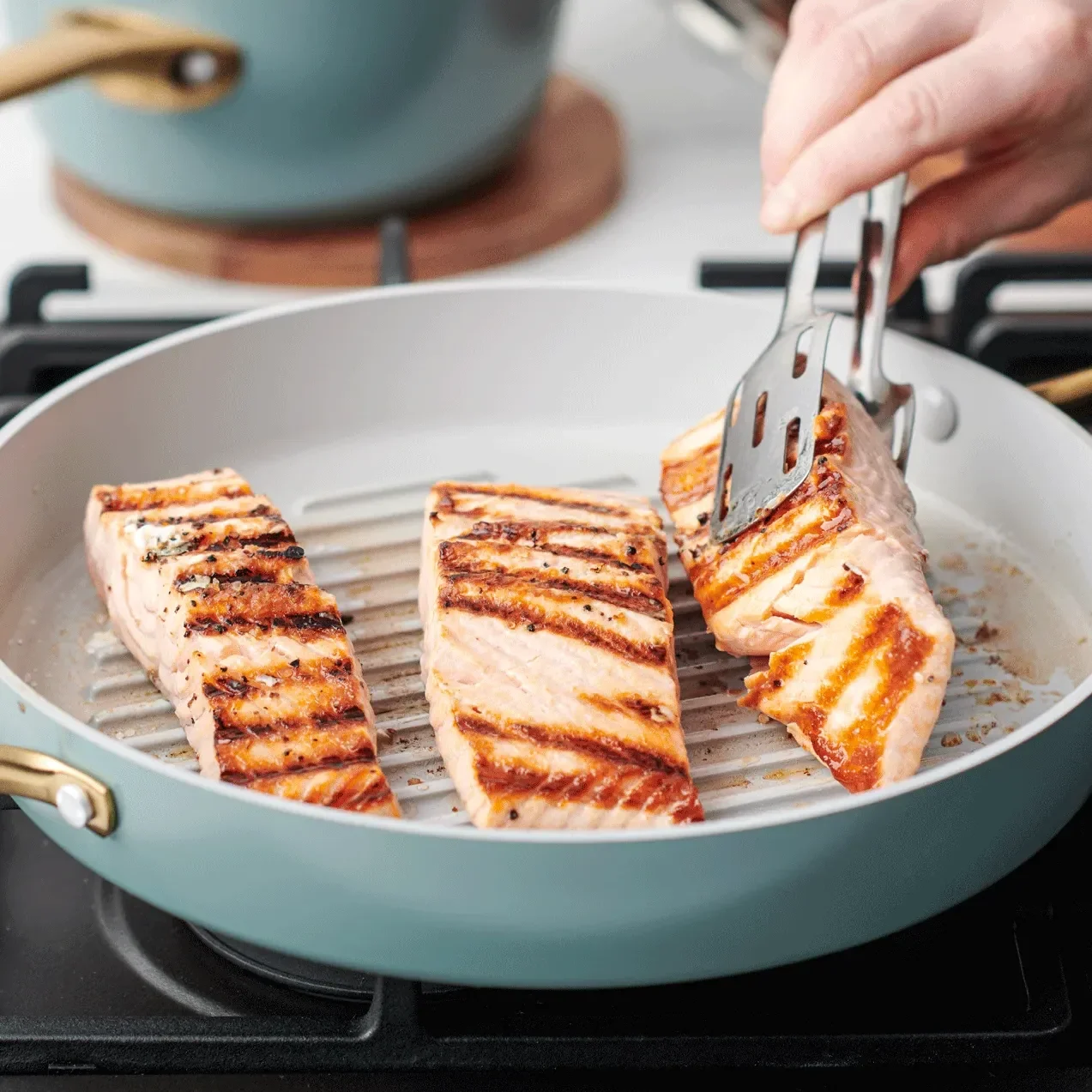 In einer blauen Pfanne von Greenpan werden drei Stücke Lachs gebraten und eines wird soeben von einer Zange gewendet.