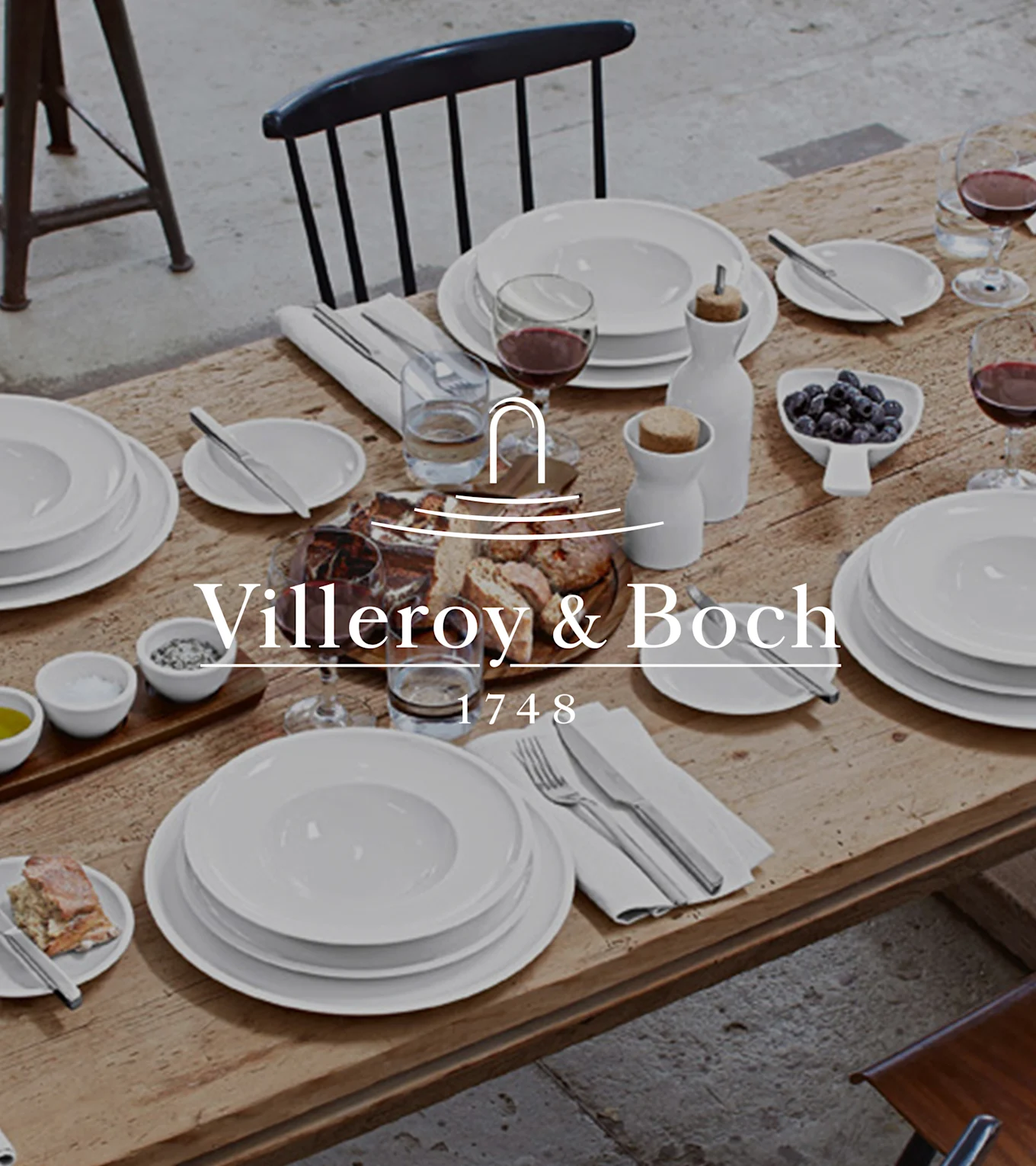 Ein rustikaler Holztisch, gedeckt mit Villeroy & Boch weißem Geschirr, Wein, Brot, Heidelbeeren, Salz, Pfeffer und Olivenöl für eine Mahlzeit.