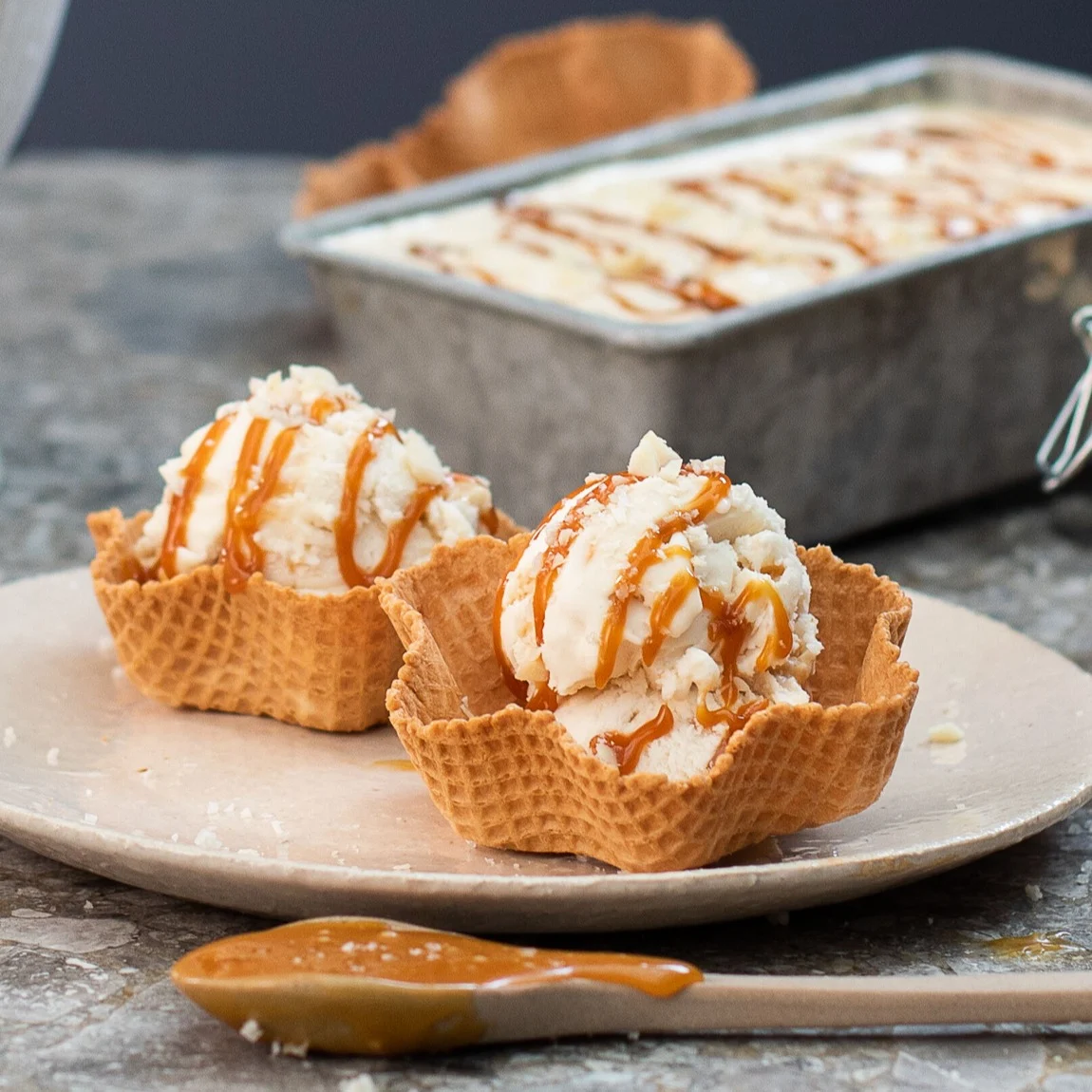 Zwei Kugeln Eis mit Karamellsauce in Waffelschalen, mit einer Form Eis im Hintergrund.