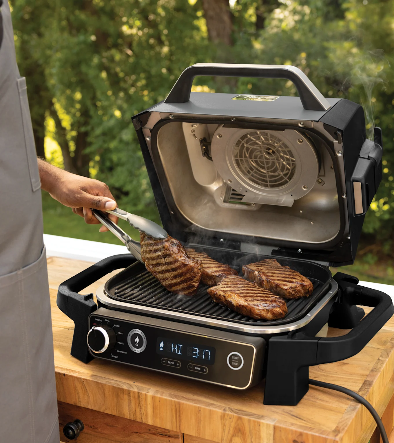 Eine Person wendet ein Steak mit einer Zange auf einem schwarzen Elektrogrill im Freien, während andere Steaks braten und Rauch aufsteigt.