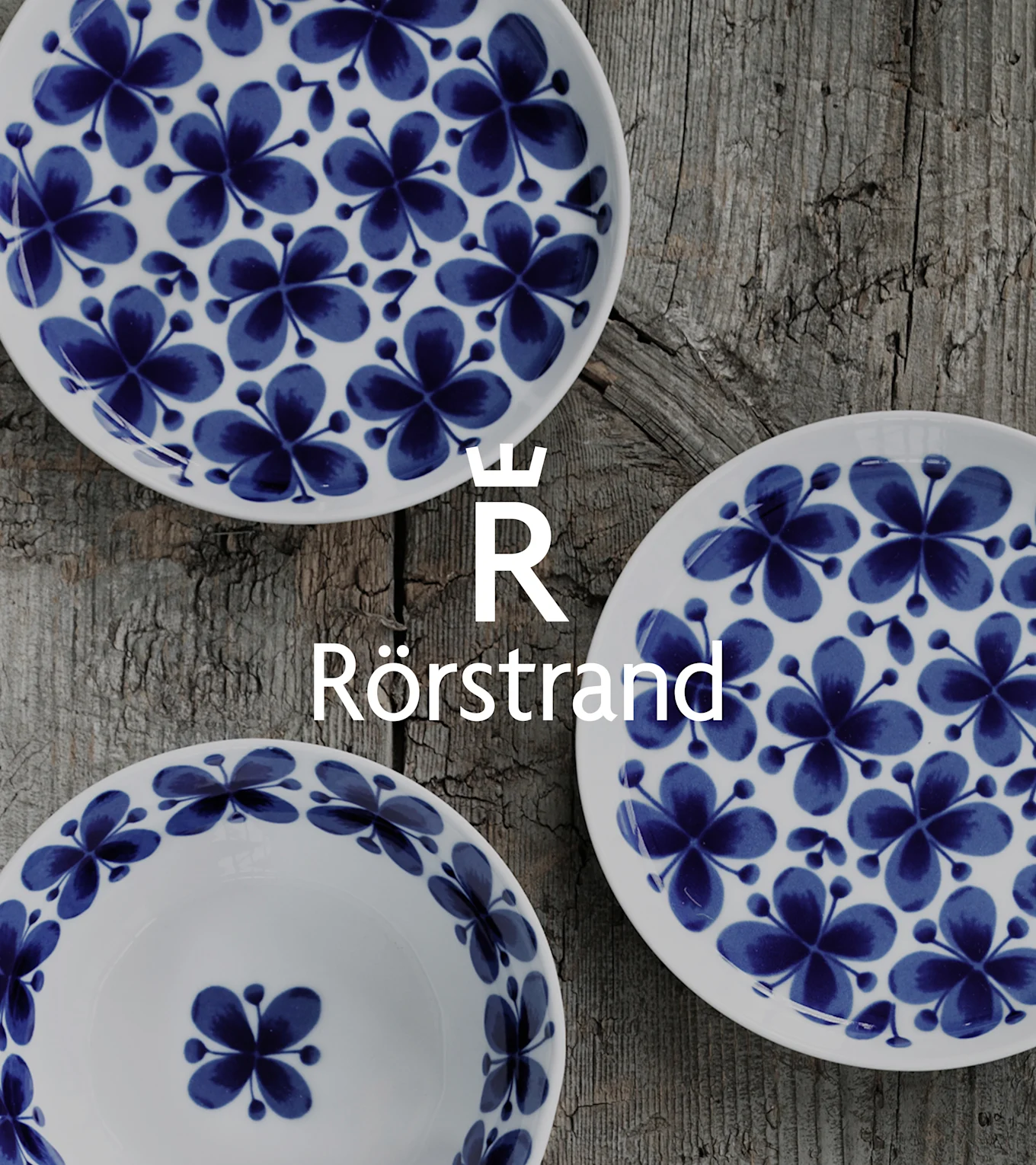 Drei weiße Teller mit blauen Blumenmustern, auf einem verwitterten Holztisch angeordnet. Rörstrand-Logo überlagert.