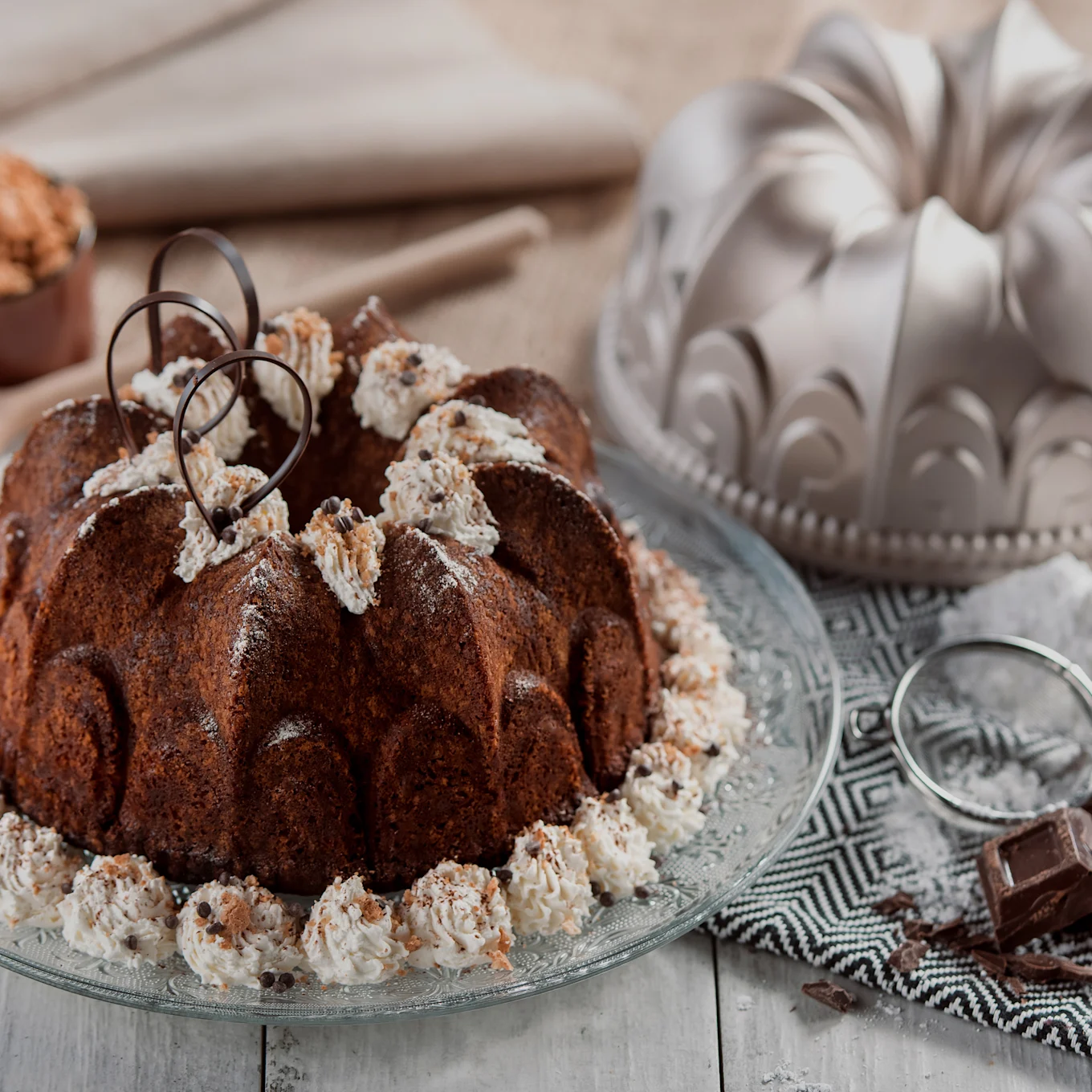 Schokoladen-Gugelhupf mit Schlagsahne, Schokoladenstückchen und Schokoladenlocken auf einer Glasplatte, mit einer Gugelhupfform und Zutaten im Hintergrund.