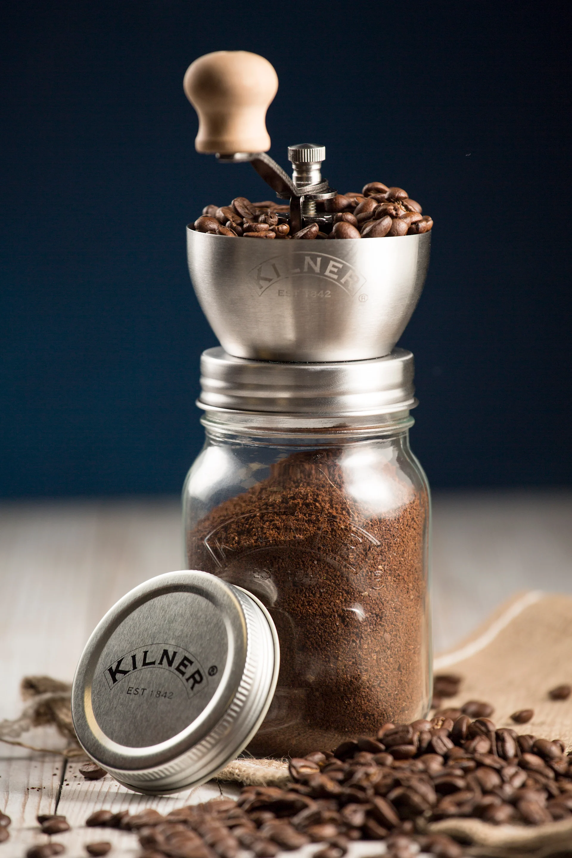 Manuelle Kaffeemühle mit ganzen Bohnen auf einem Glas gemahlenem Kaffee, Deckel daneben, verstreute Bohnen auf Holz.