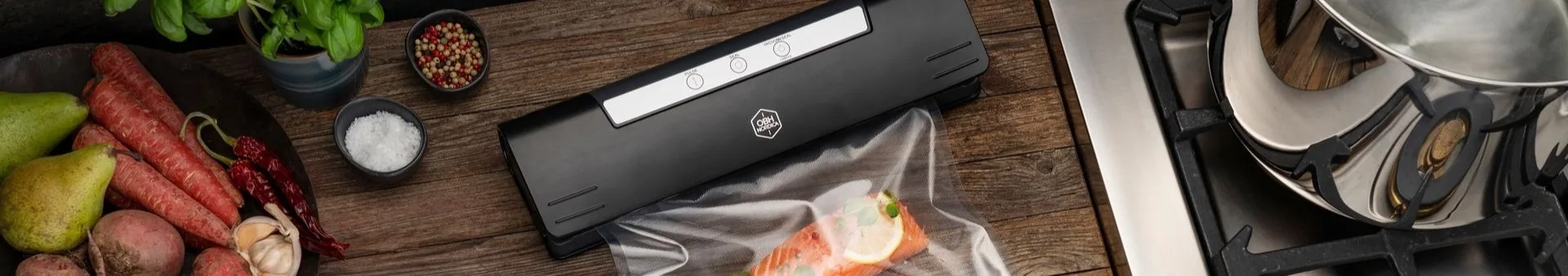 Schwarzer Vakuumierer verpackt Lachs, umgeben von frischem Gemüse, Kräutern und Gewürzen auf einer Holztheke.