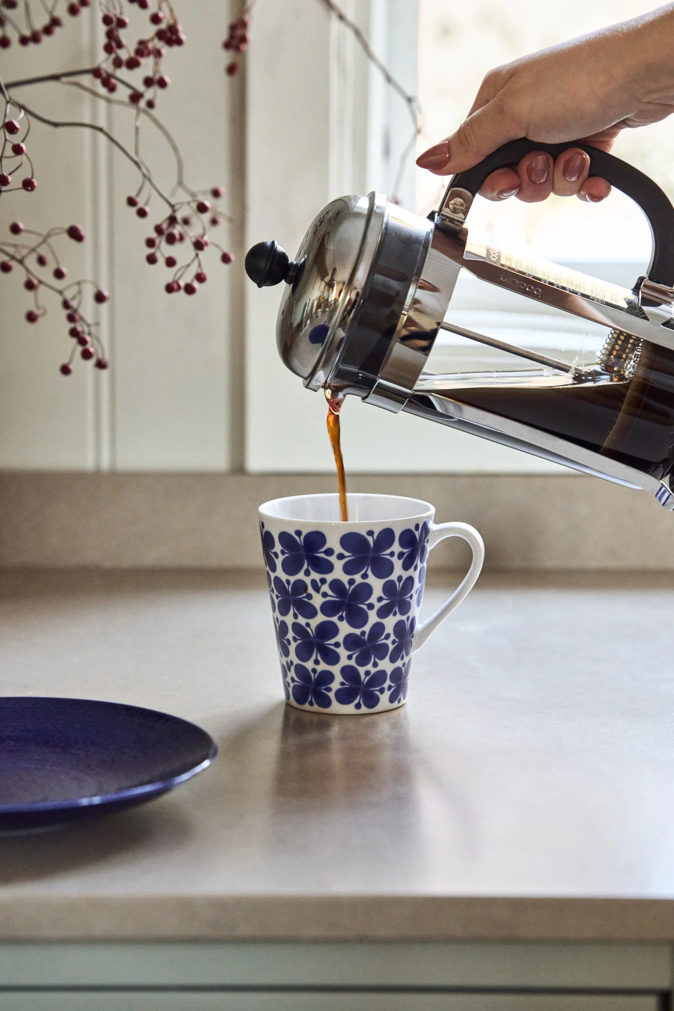 Hand gießt Kaffee aus einer French Press in eine weiße Tasse mit blauem Blumenmuster auf einer Arbeitsplatte.