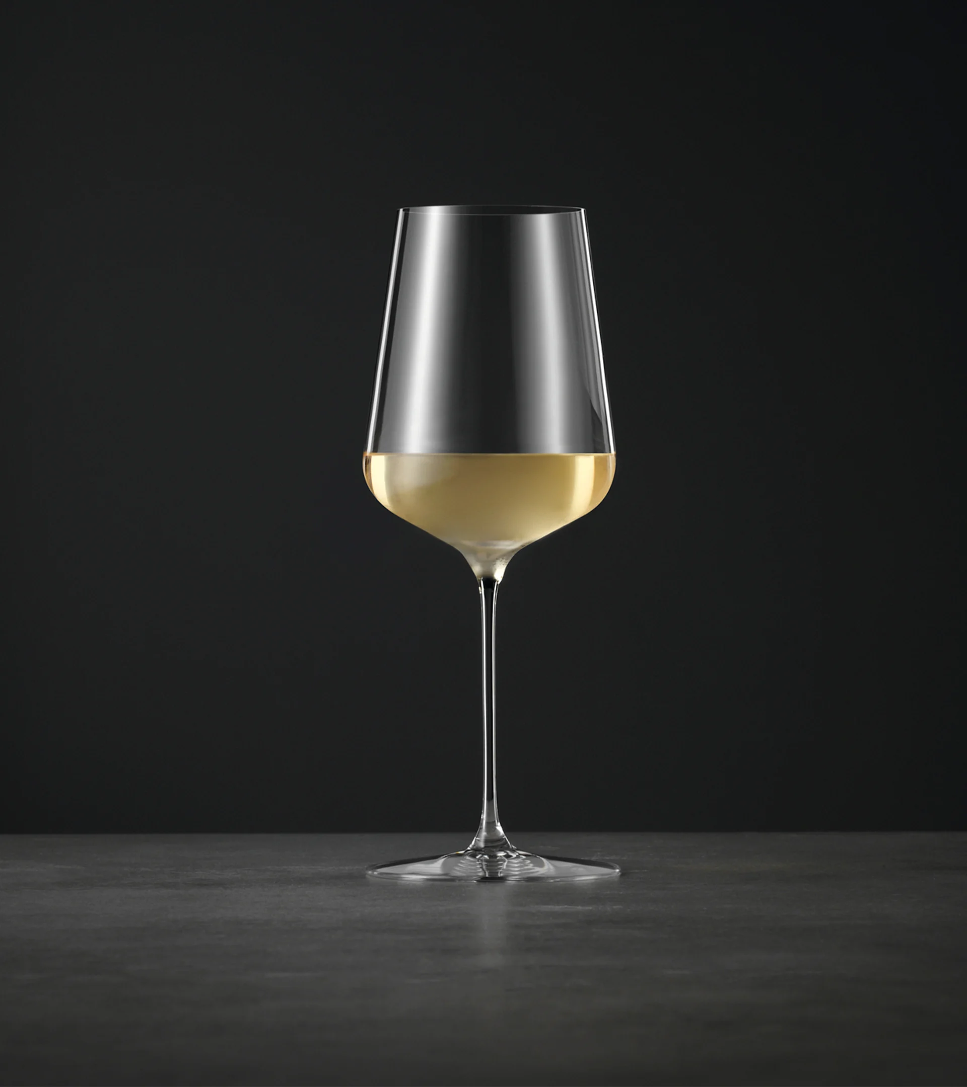 Ein hohes, elegantes Weinglas mit langem Stiel, halb gefüllt mit hellem Weißwein, vor dunklem Hintergrund.