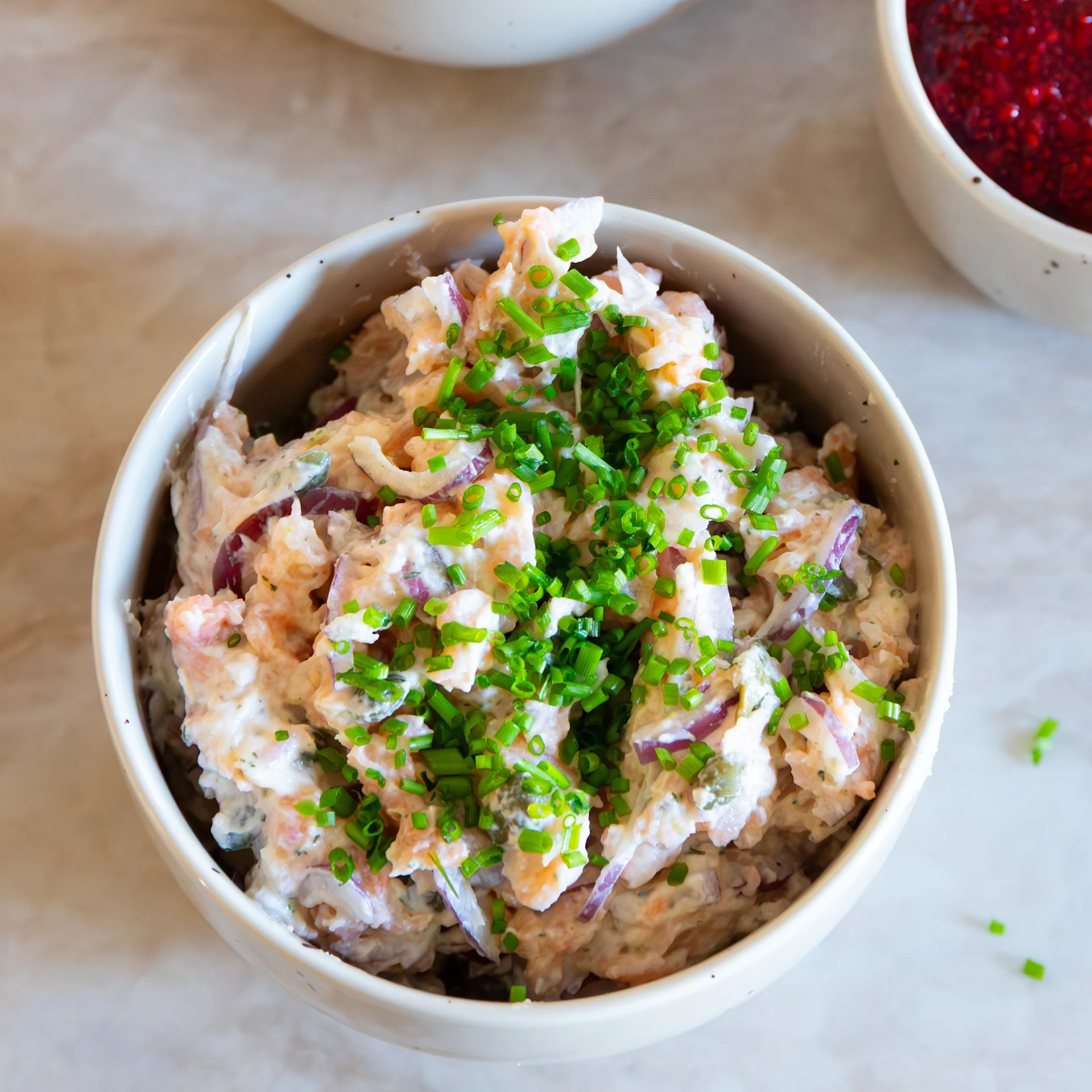 Schüssel mit cremiger Garnelensalat mit roten Zwiebeln und Schnittlauch, mit einer teilweise sichtbaren Schüssel mit roten Beeren im Hintergrund.