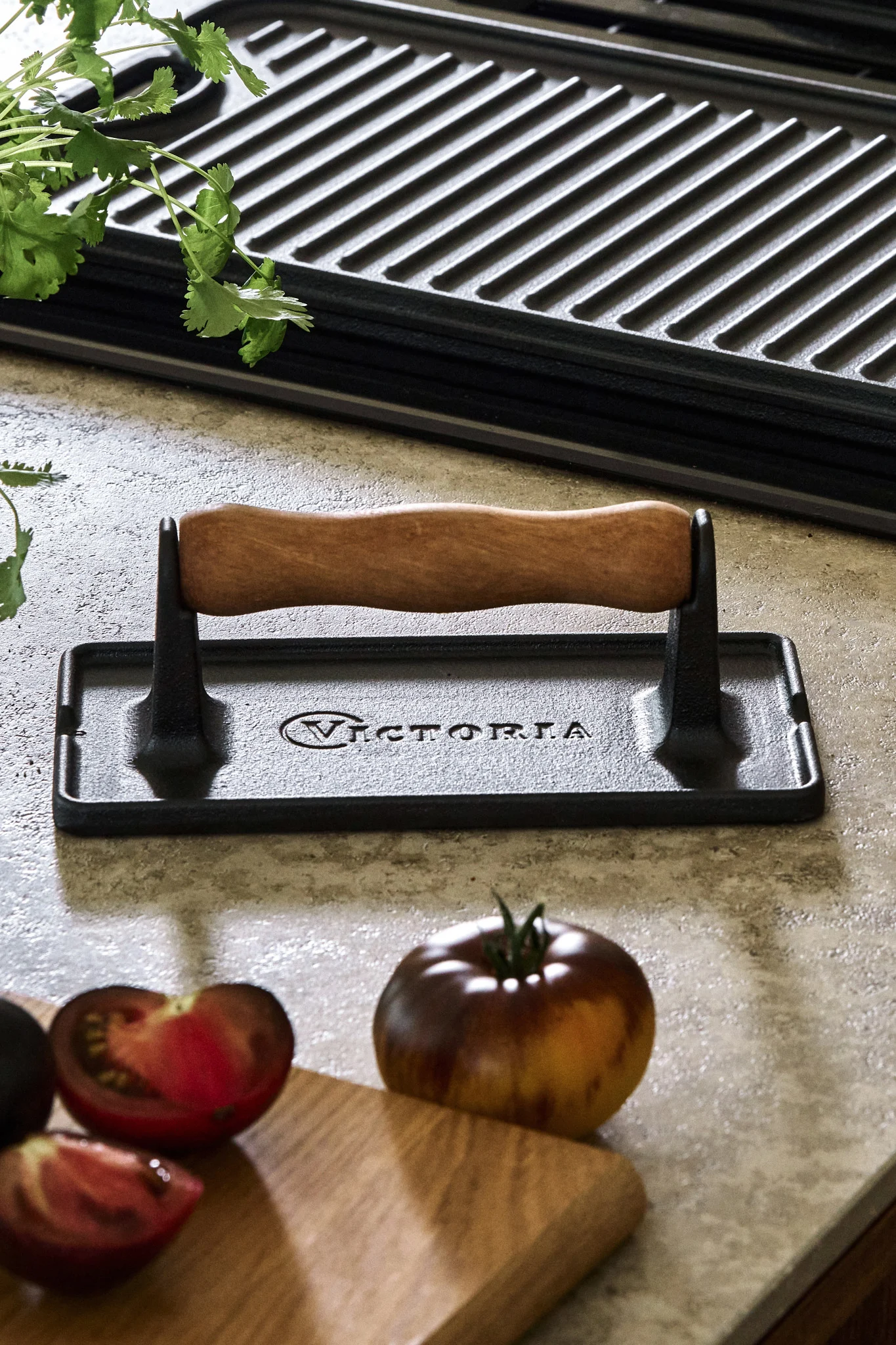 Eine Fleischpresse aus Gusseisen, die neben Tomaten und einer Grillplatte auf einer Küchenablage steht. 