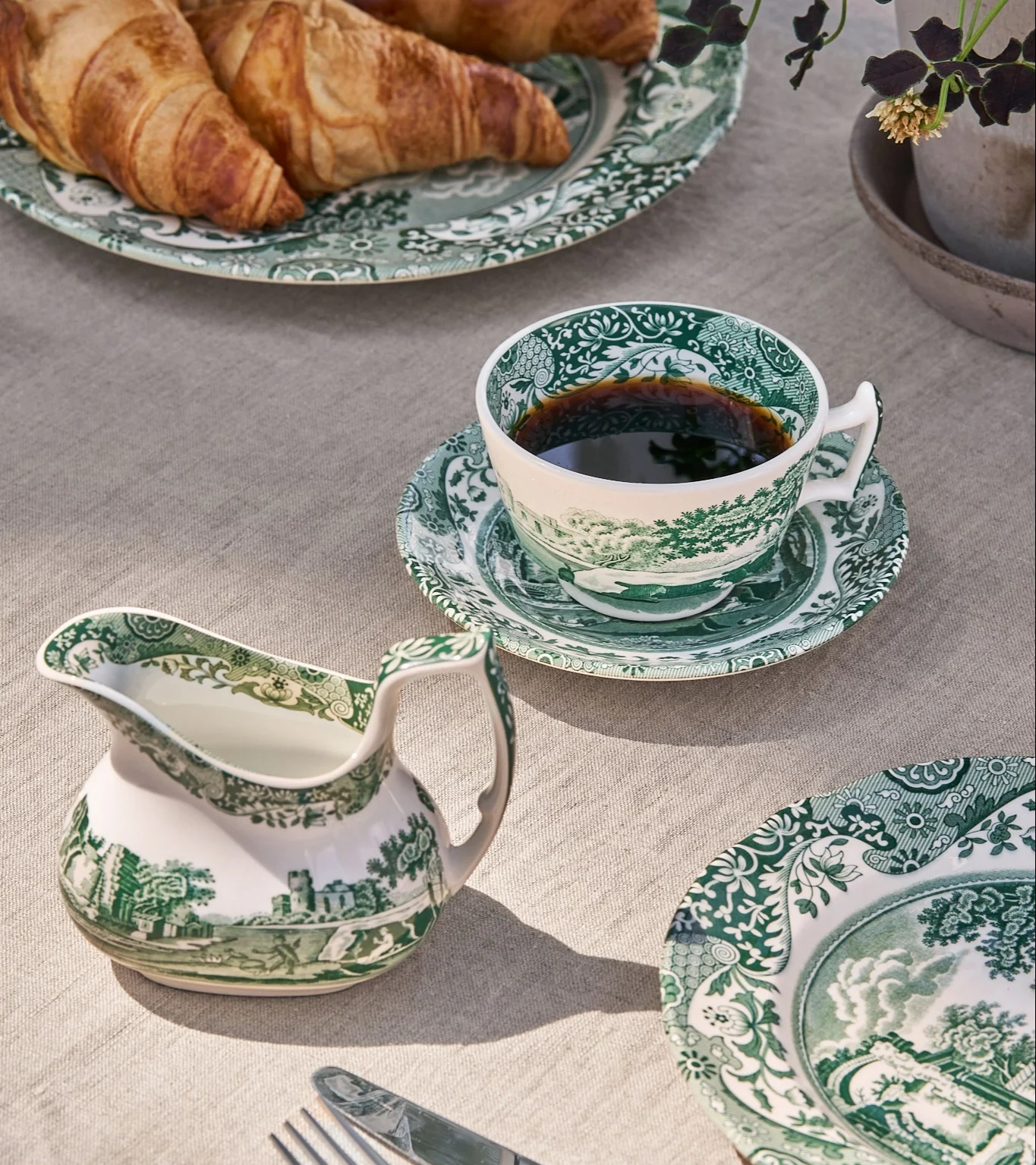 Frühstückstisch mit grün-weiß gemusterter Porzellantasse mit Kaffee, Milchkännchen und Tellern mit Croissants.