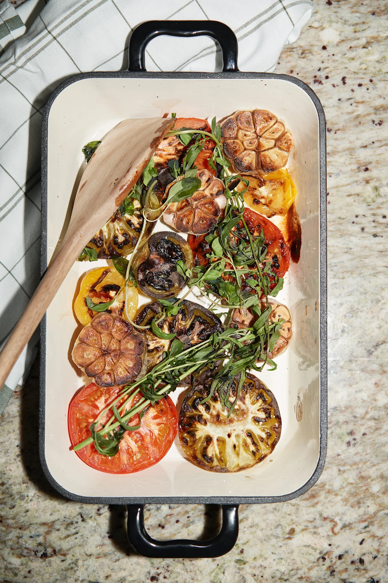 Gegrillte Tomaten, Knoblauch und frisches Grün in einer weißen Auflaufform mit Holzlöffel auf einer Granitarbeitsplatte.