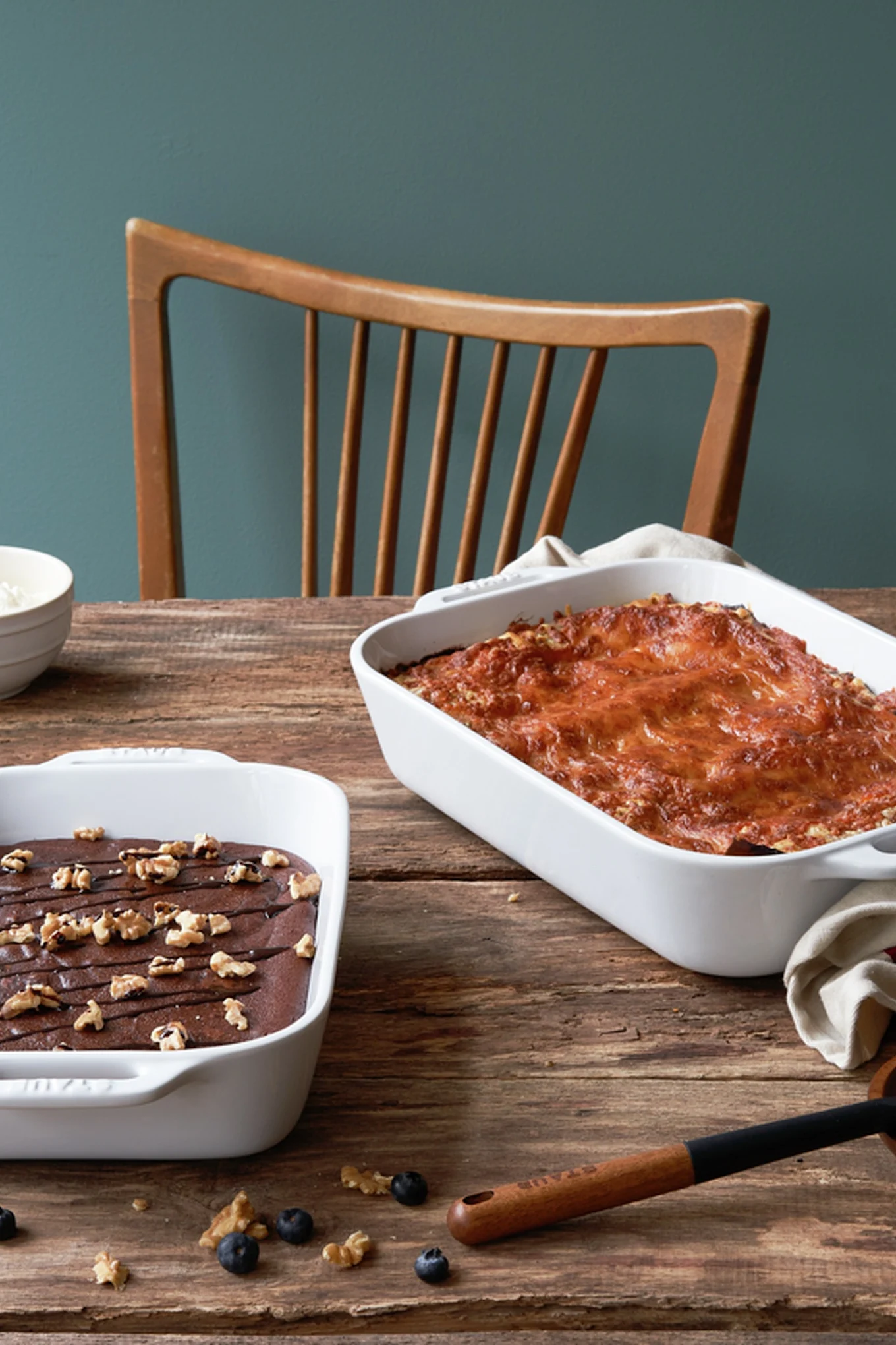 Zwei Auflaufformen von Staub, eine kleinere mit Schokokuchen und eine größere mit Lasagne, stehen auf einem Holztisch.
