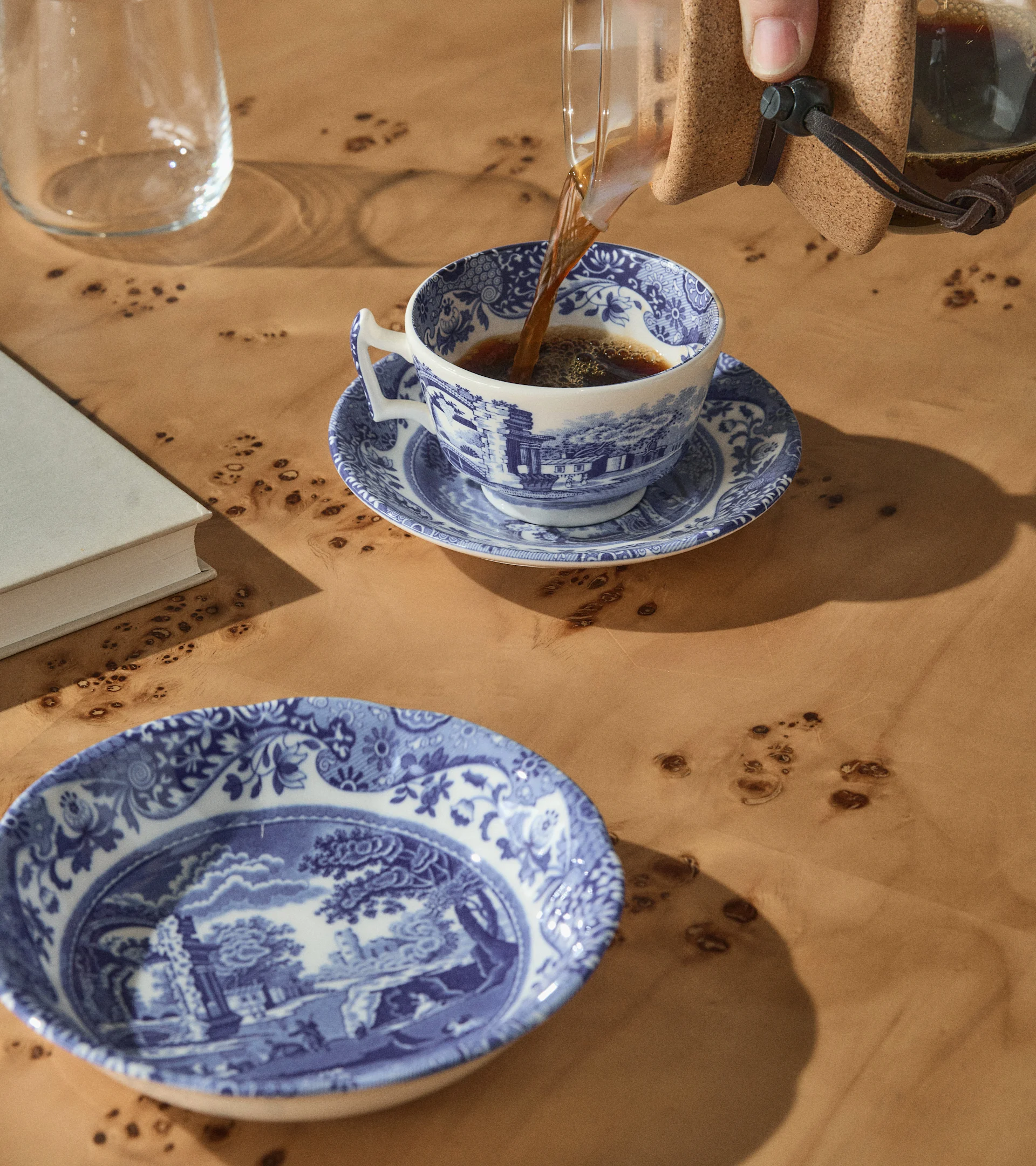 Eine Hand gießt Kaffee in eine blau-weiß gemusterte Teetasse auf einem Holztisch mit passender Schale, einem Buch und einem Glas.