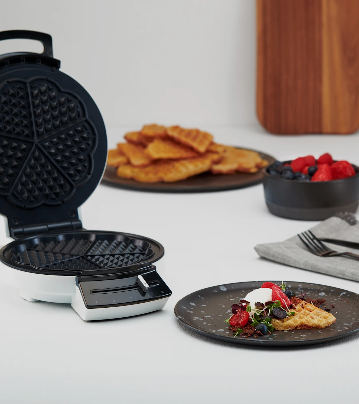 Offenes Waffeleisen neben Teller mit Waffel, Schlagsahne, Beeren und Mikrogrün. Weitere Waffeln und Beeren im Hintergrund.