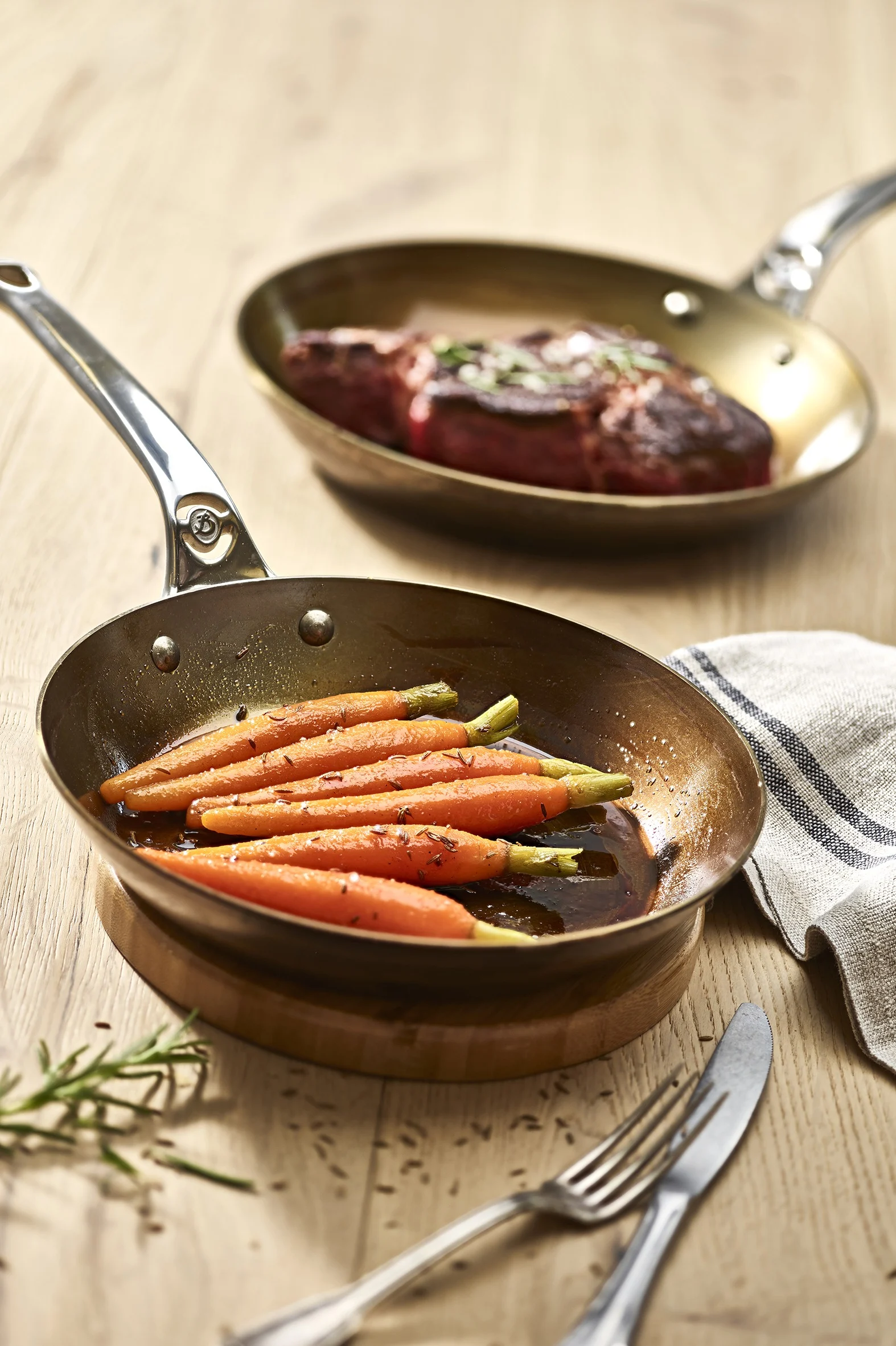 Gekochte Babykarotten in einer Kupferpfanne im Vordergrund, mit einem gebratenen Steak in einer weiteren Kupferpfanne im unscharfen Hintergrund.