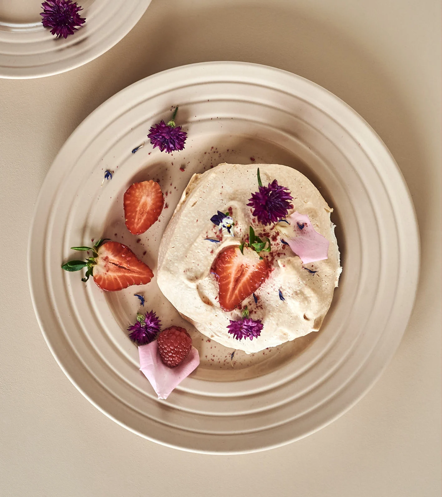 Ein cremiges Dessert mit Erdbeeren, einer Himbeere und lila Blüten auf einem beigen, geriffelten Teller.