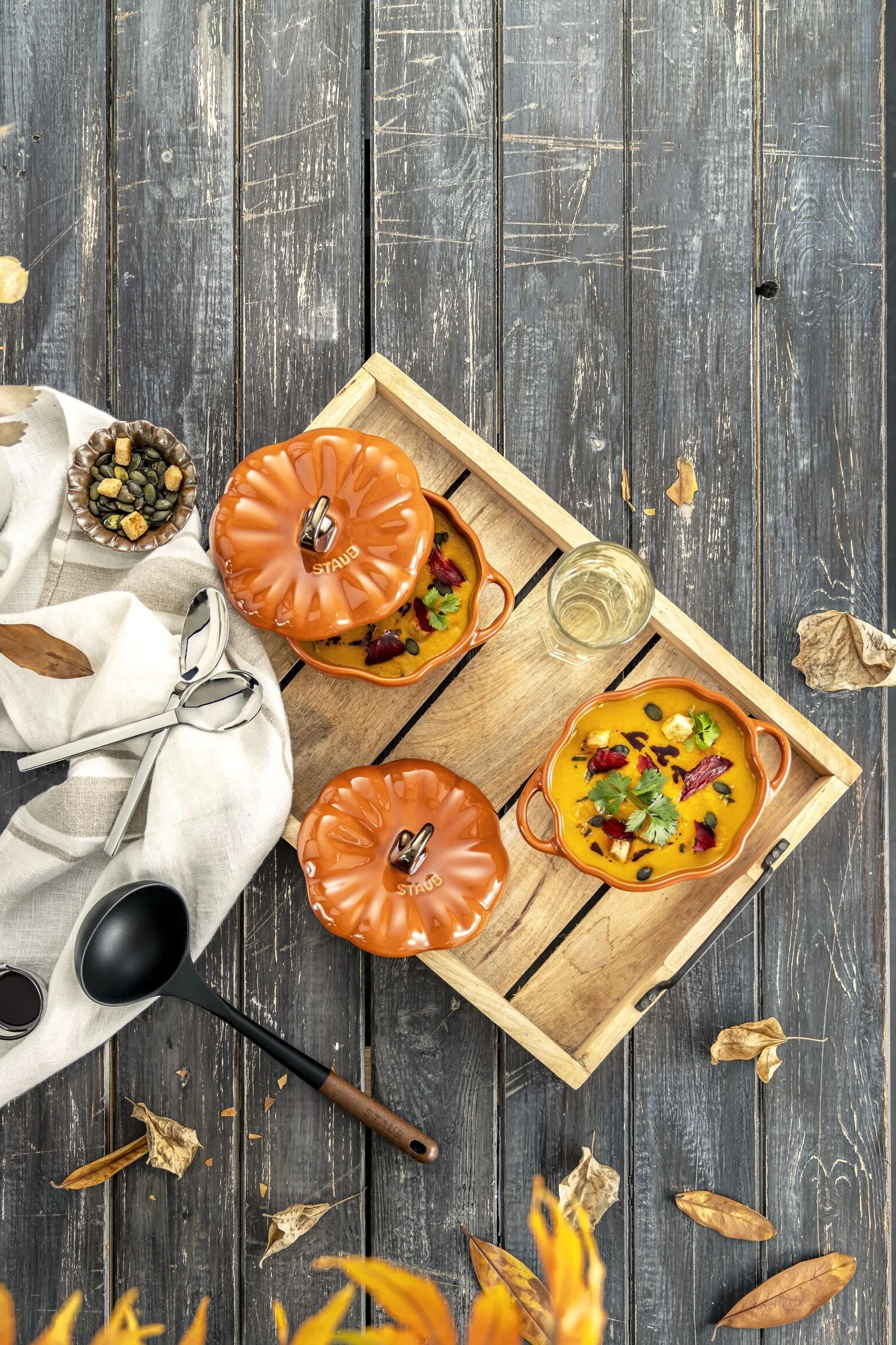Zwei orange, kürbisförmige Suppenschalen, eine abgedeckt, auf Holztablett mit Glas, Löffeln und verstreuten Blättern.