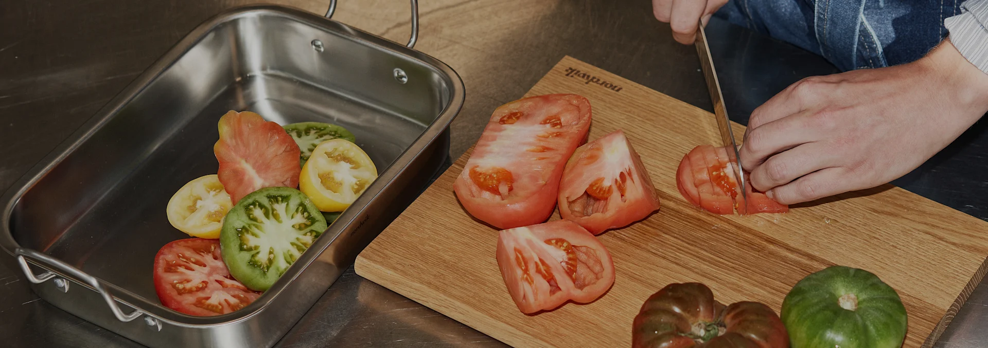 Hände schneiden Heirloom-Tomaten auf einem Holzschneidebrett, weitere Tomaten in einer Edelstahl-Bratpfanne.