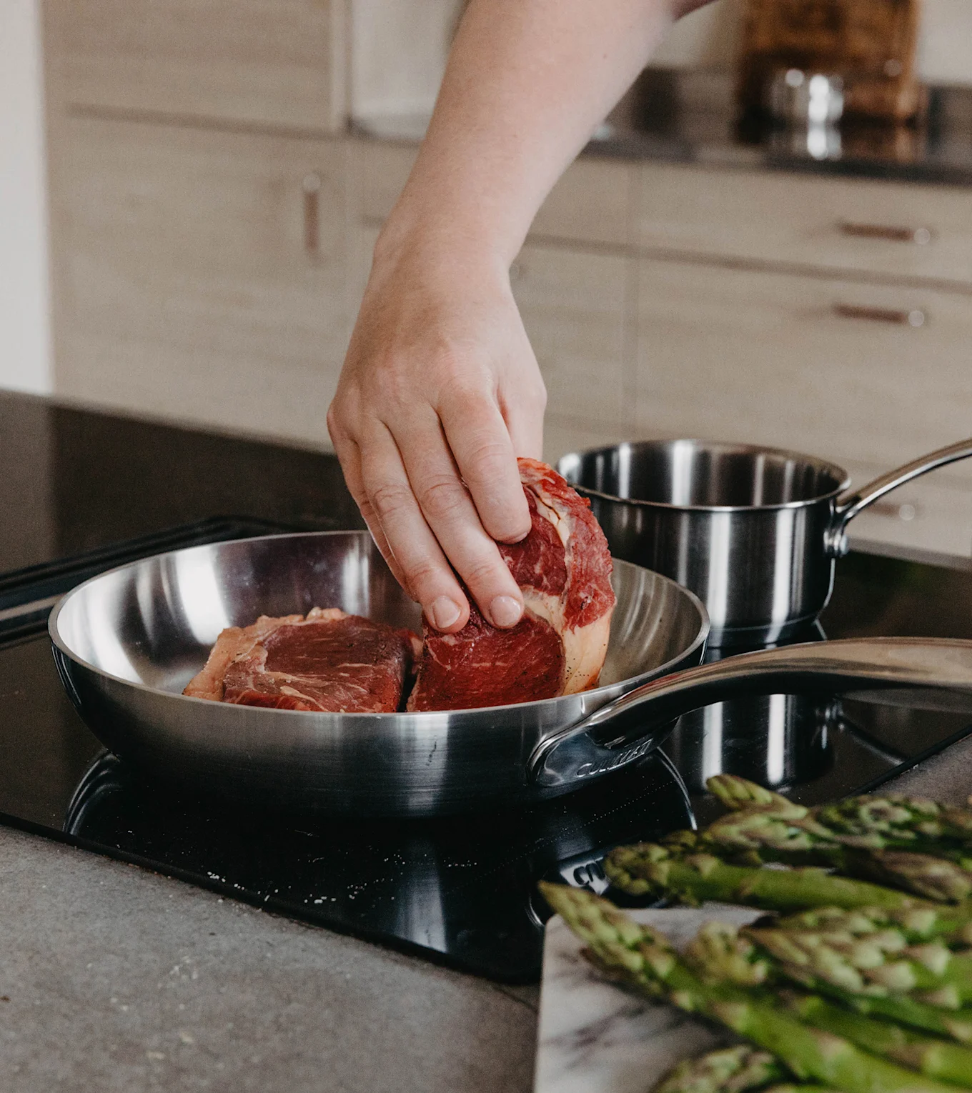Hand legt rohes Steak in Edelstahlpfanne auf Kochfeld, mit Spargel auf Marmorbrett im Vordergrund.