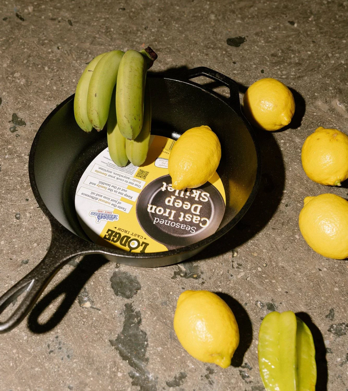 Gusseisenpfanne mit Etikett, unreifen Bananen, Zitronen und einer Sternfrucht auf dunklem Untergrund.