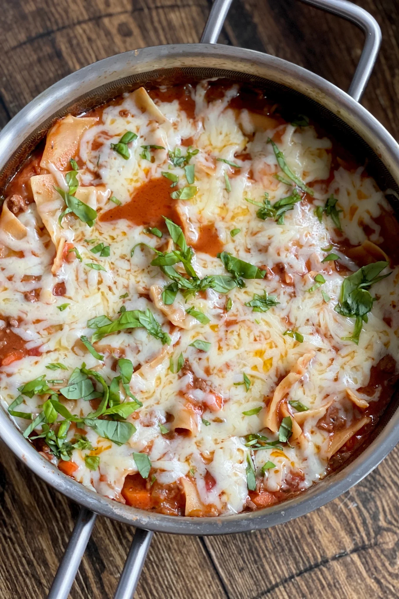 Nahaufnahme einer Pfanne mit käsiger Pastagericht mit roter Soße, Hackfleisch und frischem Basilikum auf einer Holzoberfläche.
