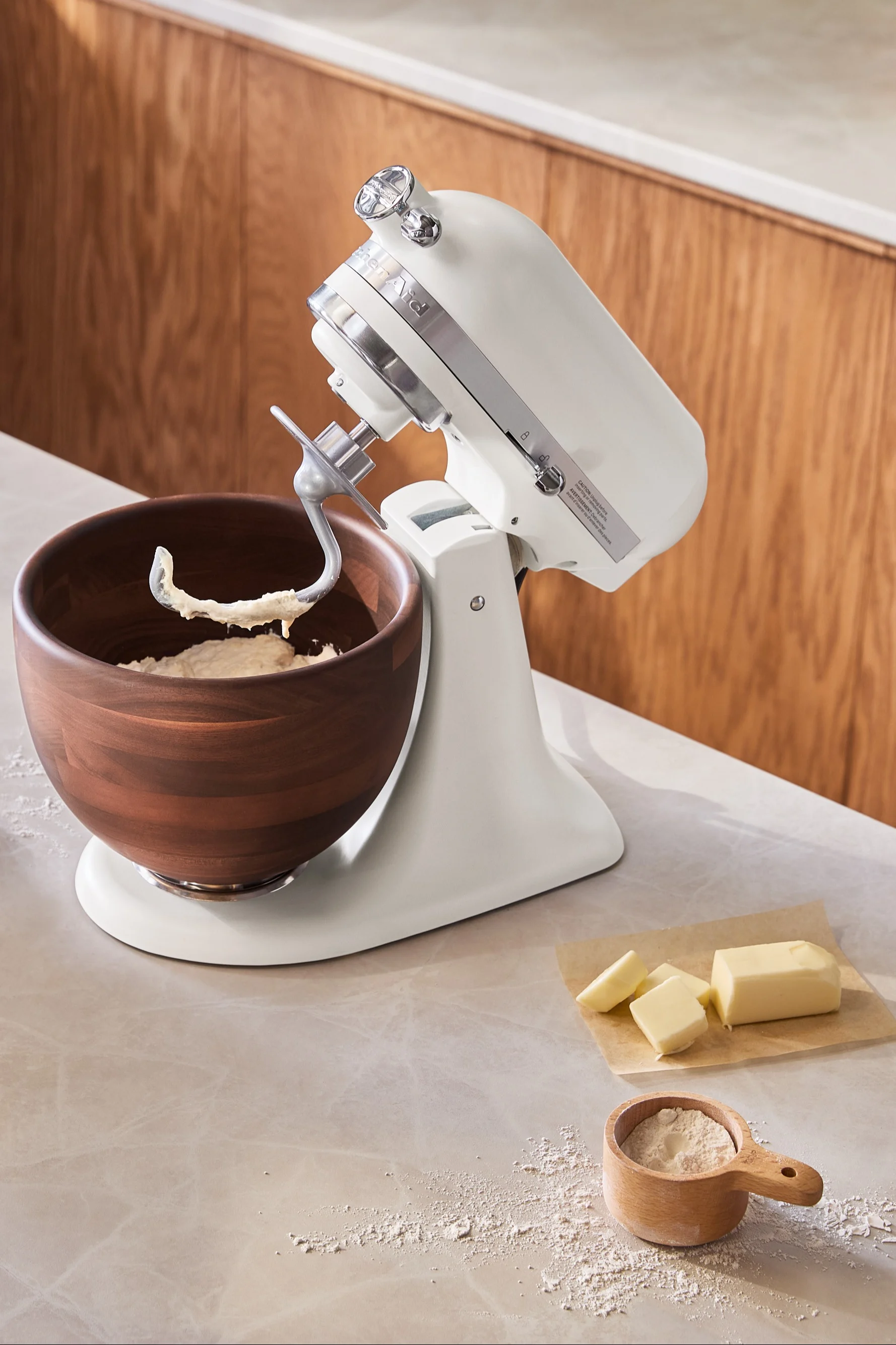 Eine weiße Küchenmaschine mit einer Holzschüssel voller Teig. Butterstücke und ein hölzerner Messbecher mit Mehl liegen auf der Arbeitsplatte.