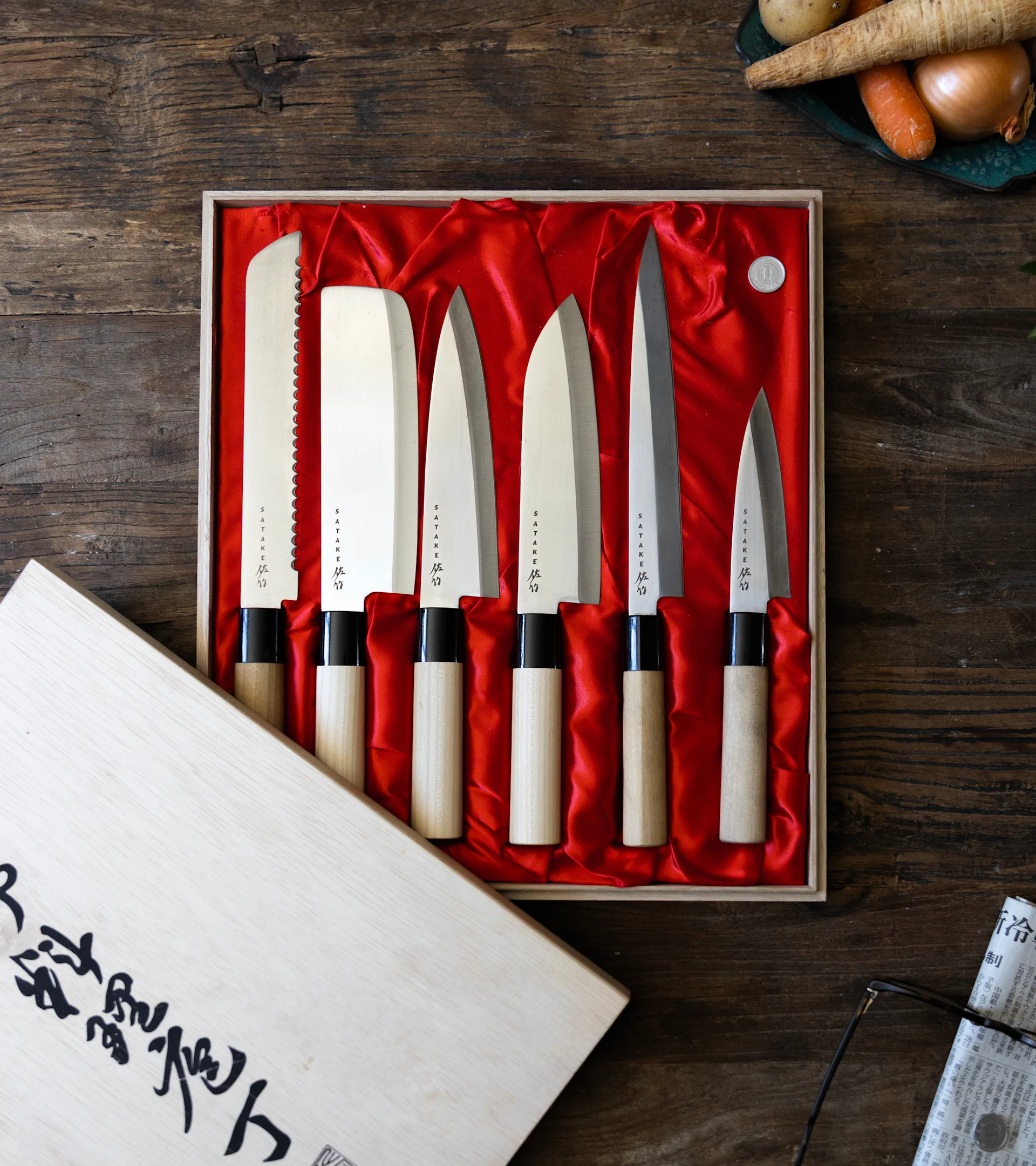 Sechs japanische Messer mit Holzgriffen in einer rot ausgekleideten Box auf einem Holztisch, mit Gemüse und Zeitung im Hintergrund.