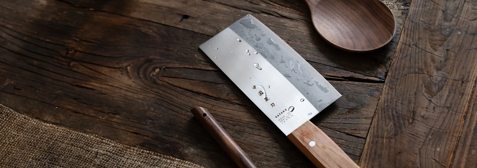 Ein Hackmesser-ähnliches Kochmesser mit Wassertropfen auf der Klinge und hellem Holzgriff, auf rustikaler Holzoberfläche mit Holzlöffel.