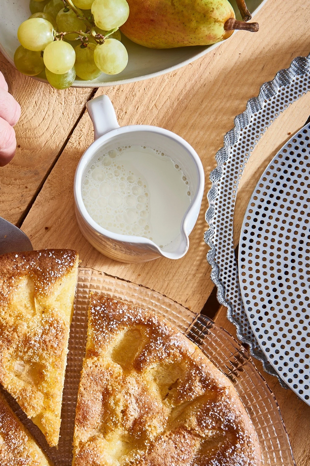 Scheiben goldener Kuchen mit Puderzucker, eine Kanne Milch, Trauben und eine Birne auf einem Holztisch.