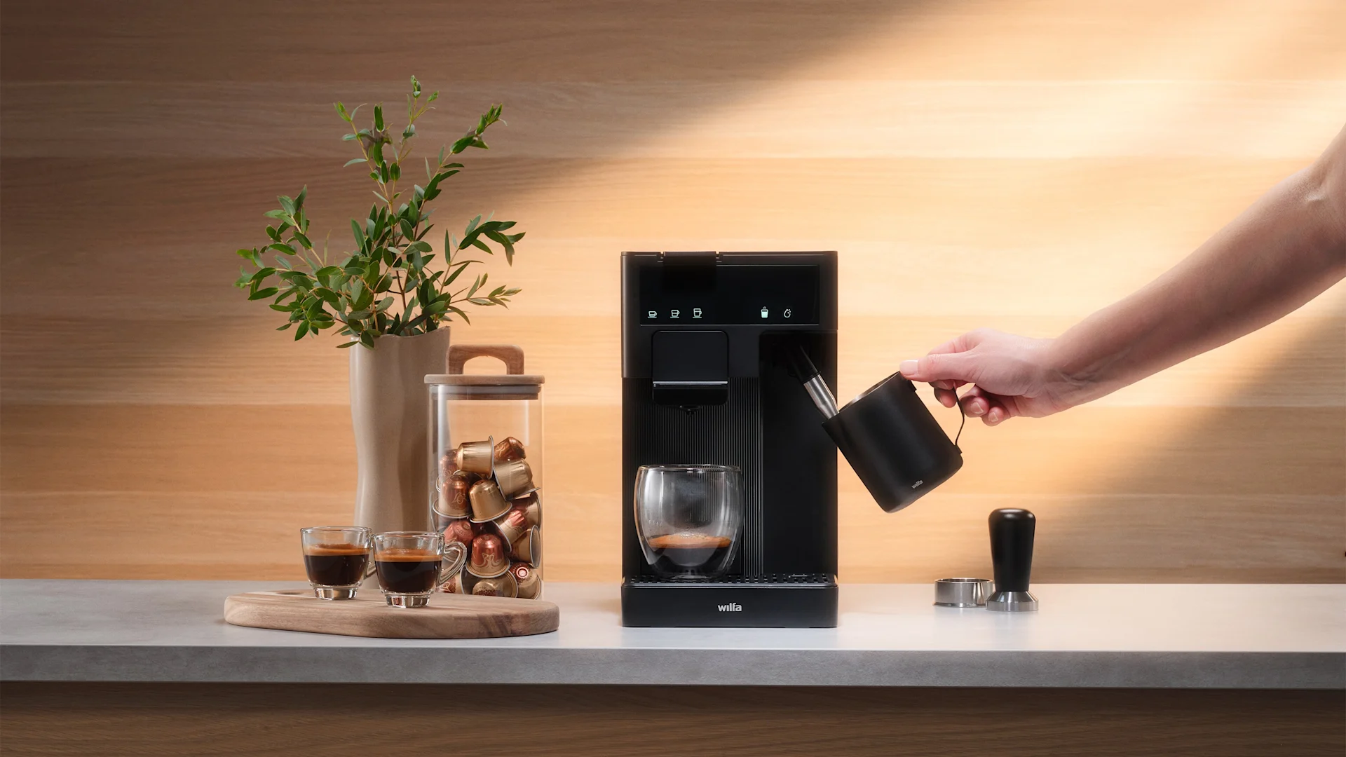 Hand dampft Milch mit schwarzem Krug und Kaffeemaschine auf Theke mit Kaffeekapseln und Espressotassen.