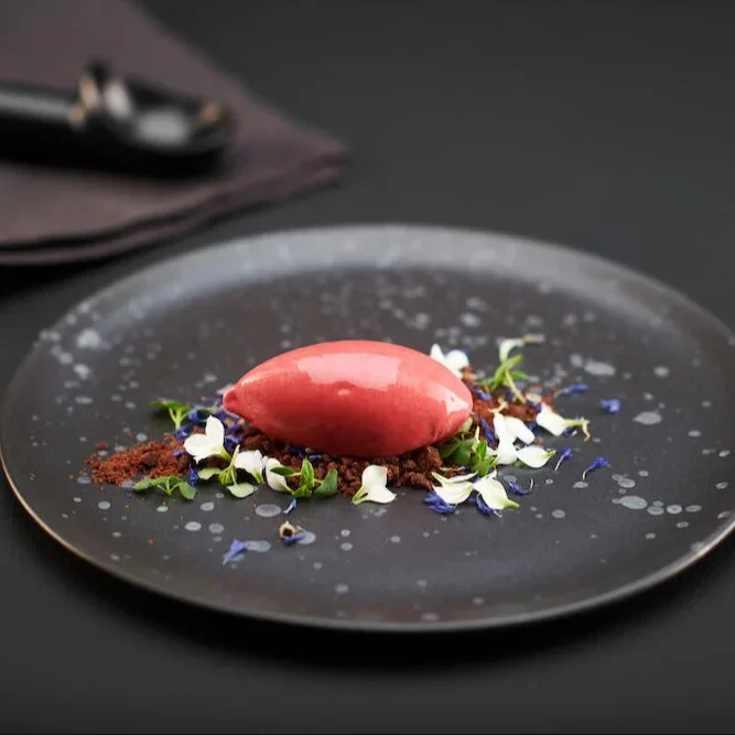 Rotes Sorbet auf Schokoladenstreuseln mit weißen und blauen Blütenblättern auf einem dunklen Teller.
