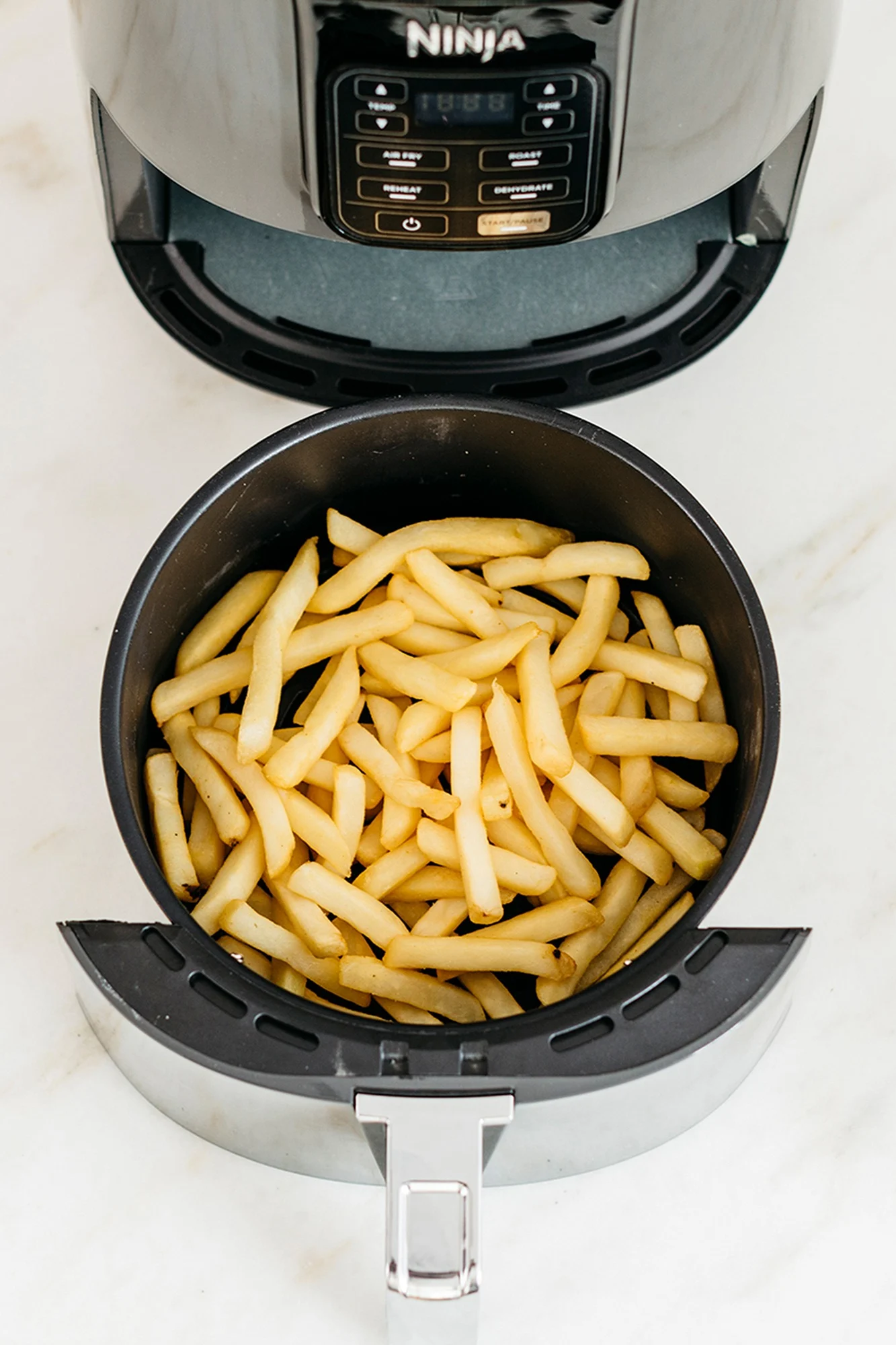 Ein Airfryer-Korb gefüllt mit goldbraunen Pommes Frites, mit dem oberen Teil eines schwarzen Airfryers darüber sichtbar.