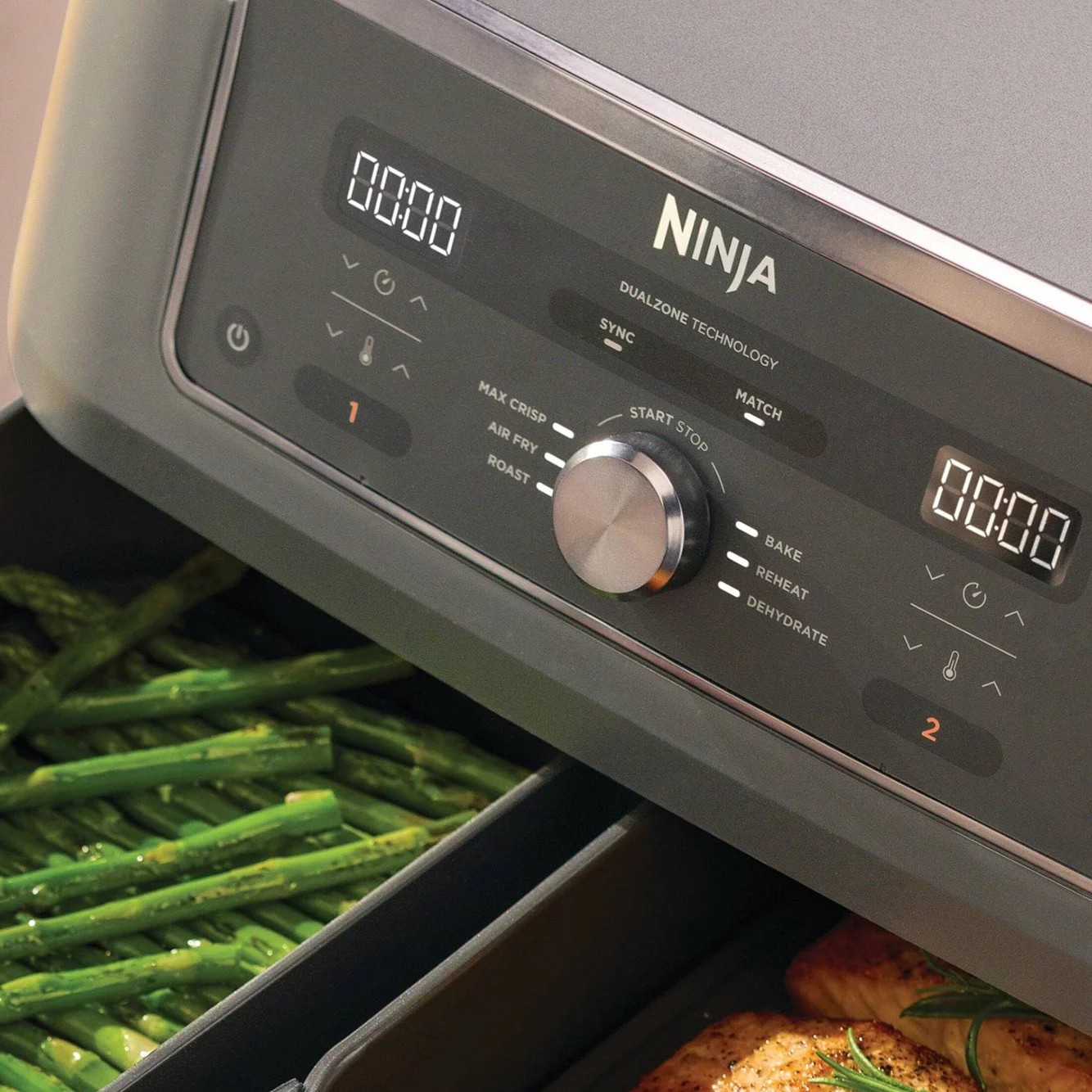 Das Bedienfeld eines Ninja Airfryers mit zwei Digitalanzeigen und einem Drehknopf, grüne Spargel in einer und Hähnchen in der anderen Schublade.