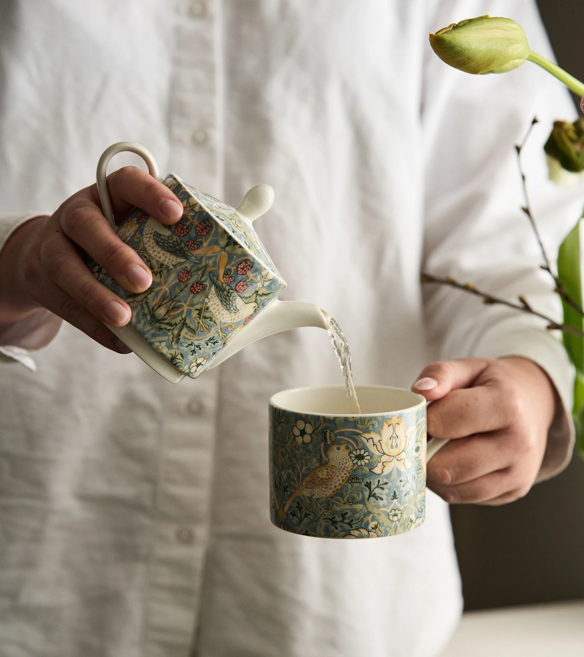 Person gießt Wasser aus einer blaugrün gemusterten Teekanne mit Vögeln und Beeren in eine passende Tasse.
