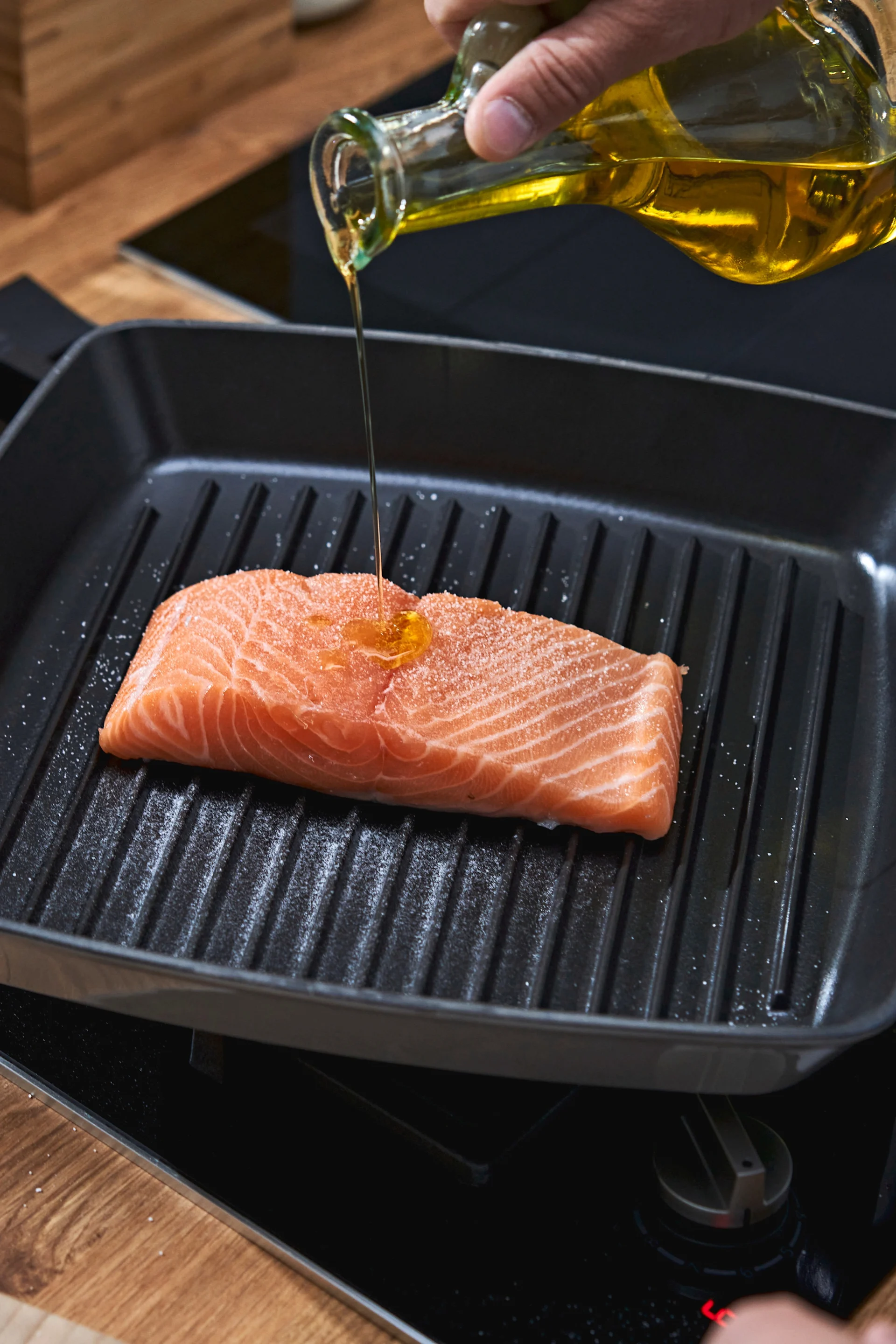 In einer Grillpfanne liegt ein Stück Lachs, der gerade mit Öl begossen wird.