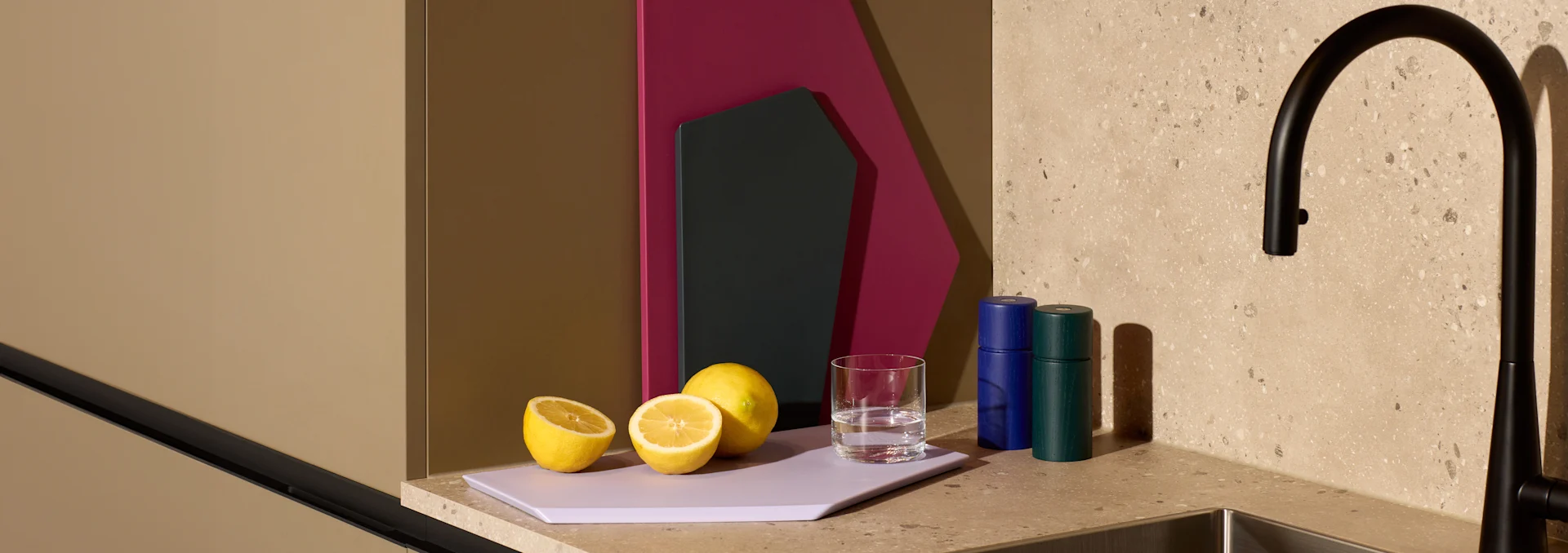 Zitronen, Wasserglas, blaue und grüne Mühlen auf lila Schneidebrett auf gesprenkelter Arbeitsplatte neben schwarzem Wasserhahn.