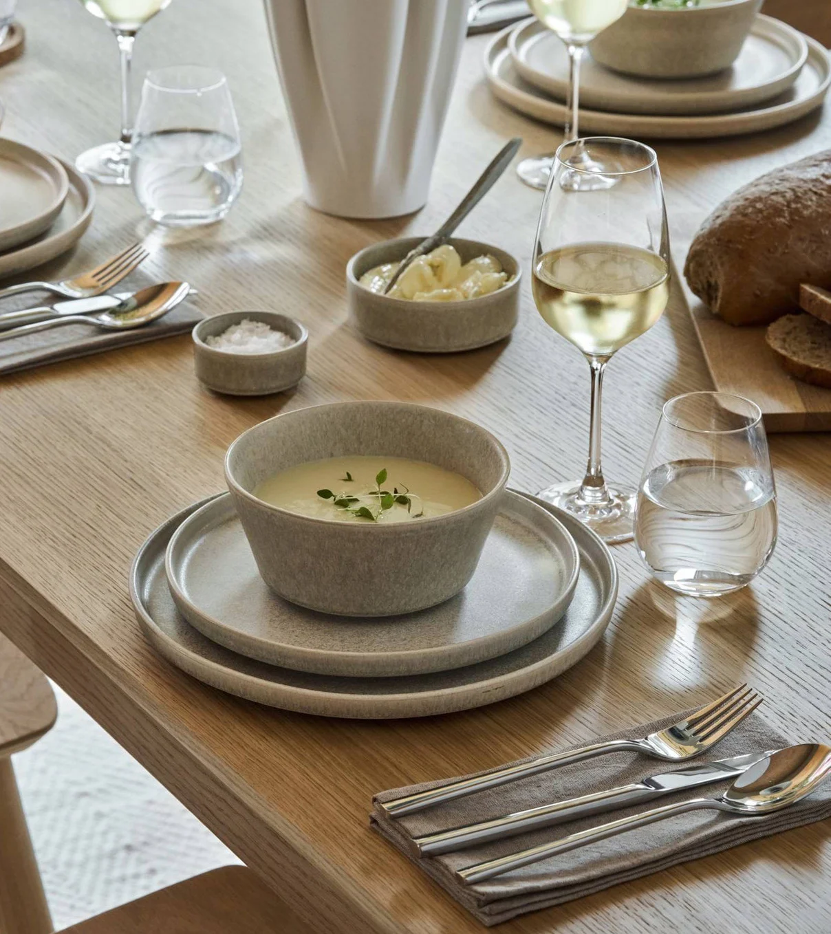 Ein Esstisch gedeckt mit hellgrauem Steingut, Suppe, Brot, Wein, Wasser und Besteck.