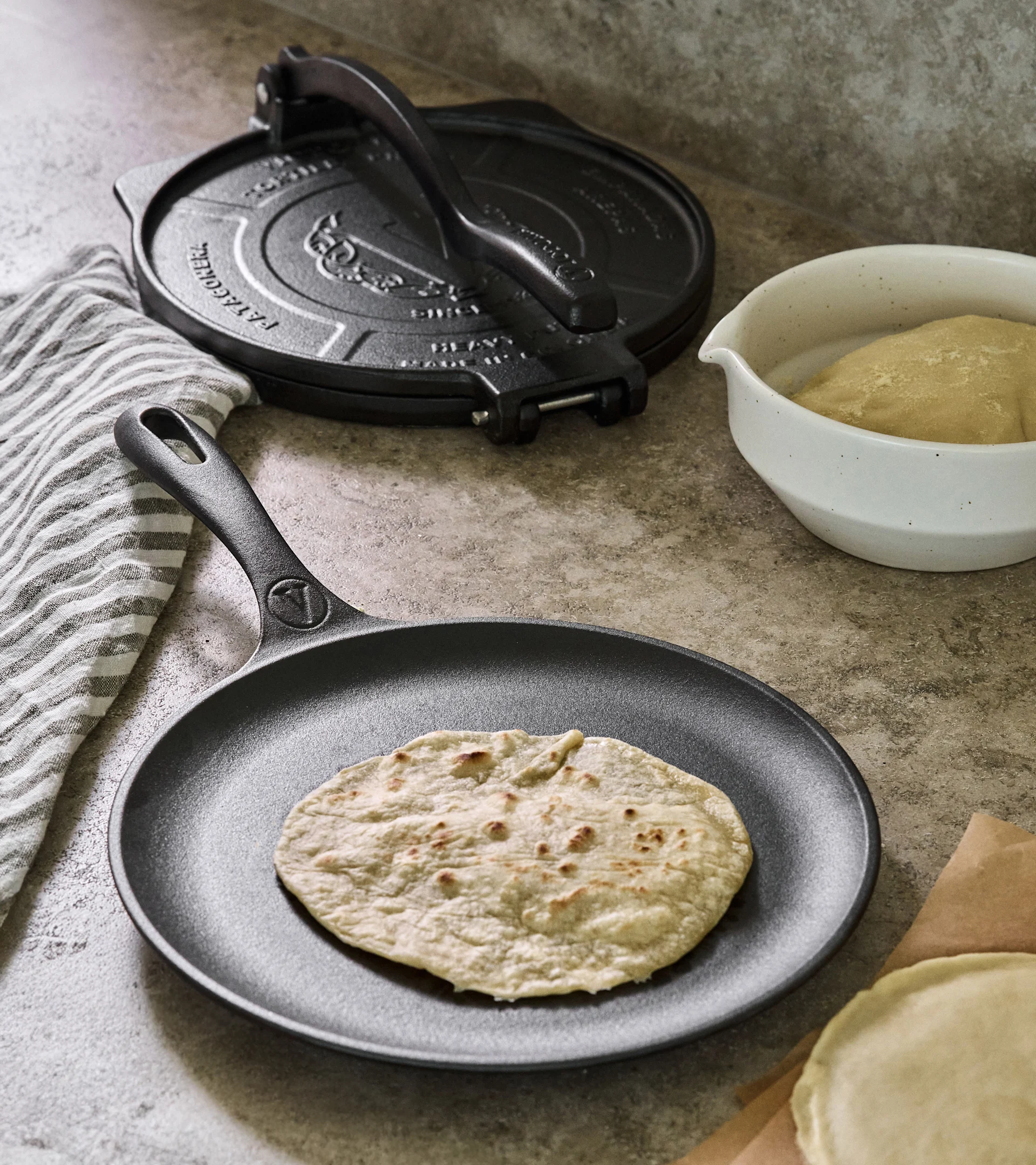Tortillapresse und Bratpfanne aus Gusseisen mit gekochter Tortilla, Teig und rohen Tortillas auf einer Theke.