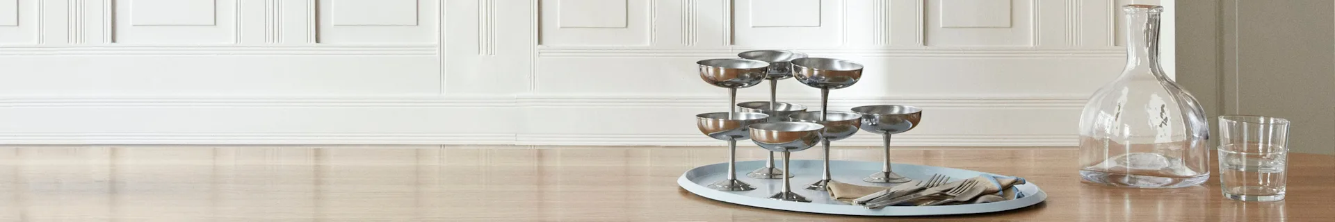 Ein hellblaues ovales Tablett mit silbernen Champagnerschalen, Besteck und Serviette auf einem Holztisch neben einer Glaskaraffe.