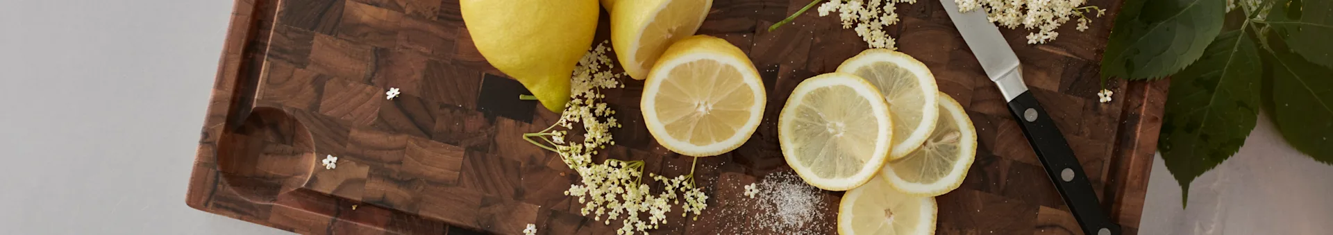 Zitronen, Holunderblüten und Zucker auf einem Holzschneidebrett mit einem Messer.
