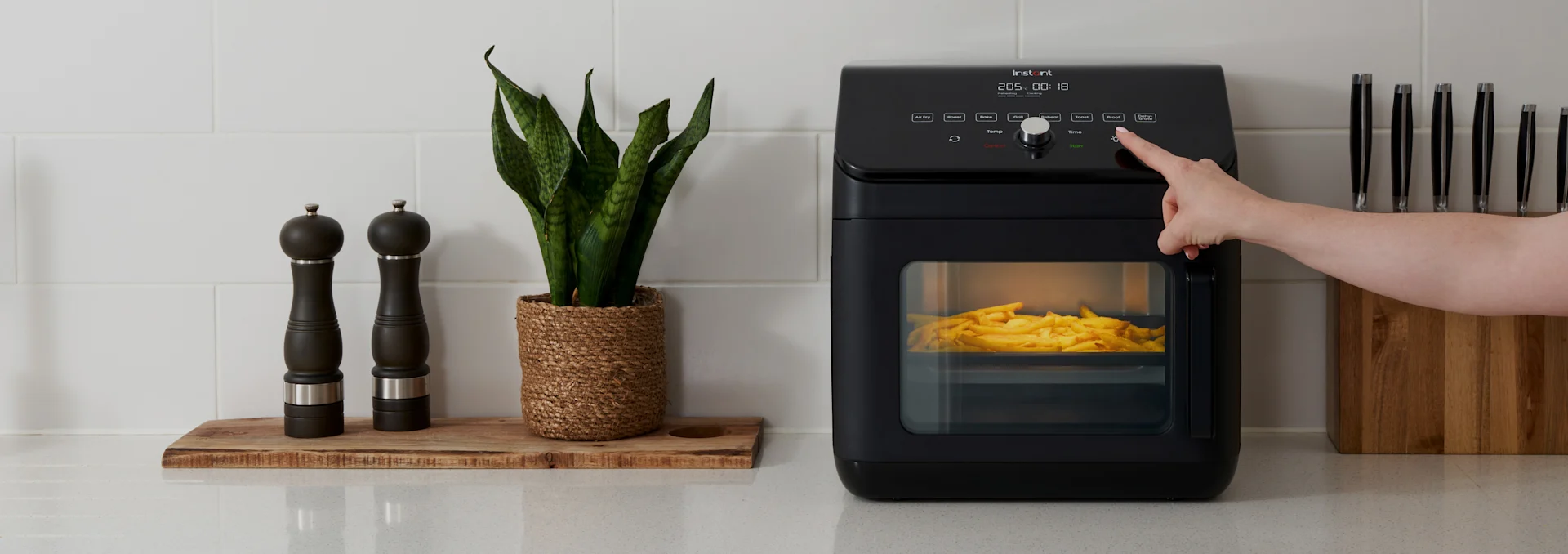 Hand drückt Taste auf schwarzem Airfryer mit Pommes frites, neben Gewürzmühlen, einer Pflanze und Messerblock auf der Arbeitsplatte.