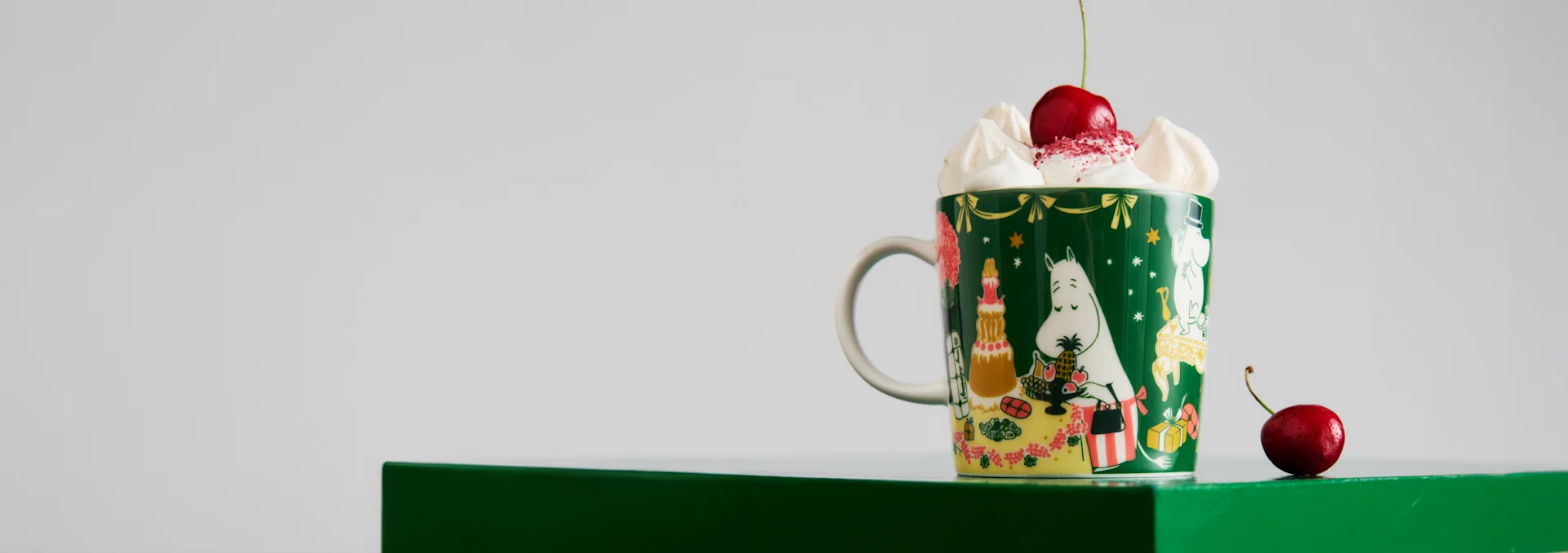 Festliche grüne Mumin-Tasse mit Schlagsahne und Kirsche, auf einer grünen Oberfläche.