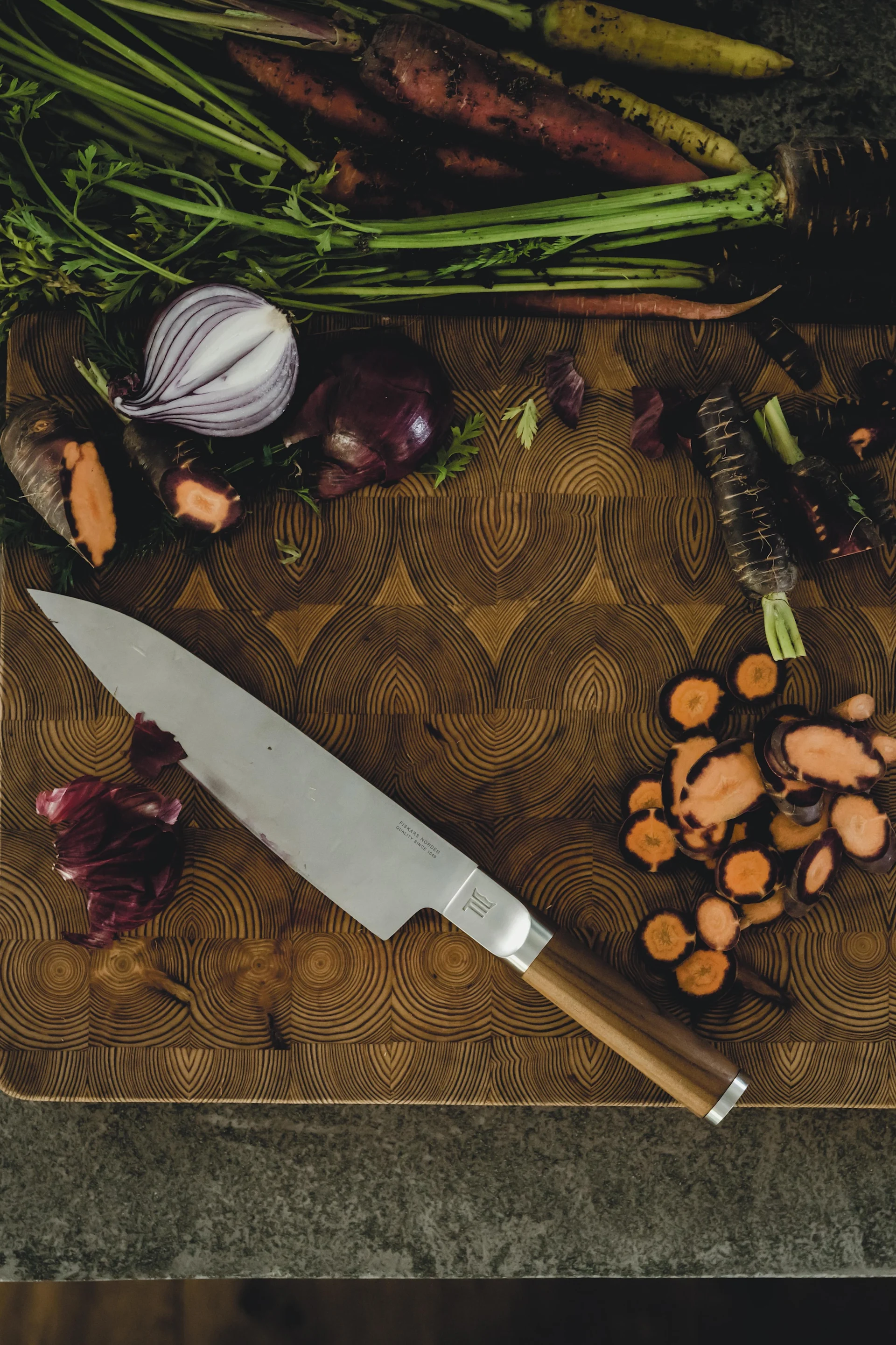 Draufsicht auf ein Küchenmesser und farbenfrohe Wurzelgemüse, darunter ganze und geschnittene lila Karotten und eine rote Zwiebel, auf einem Holzschneidebrett.