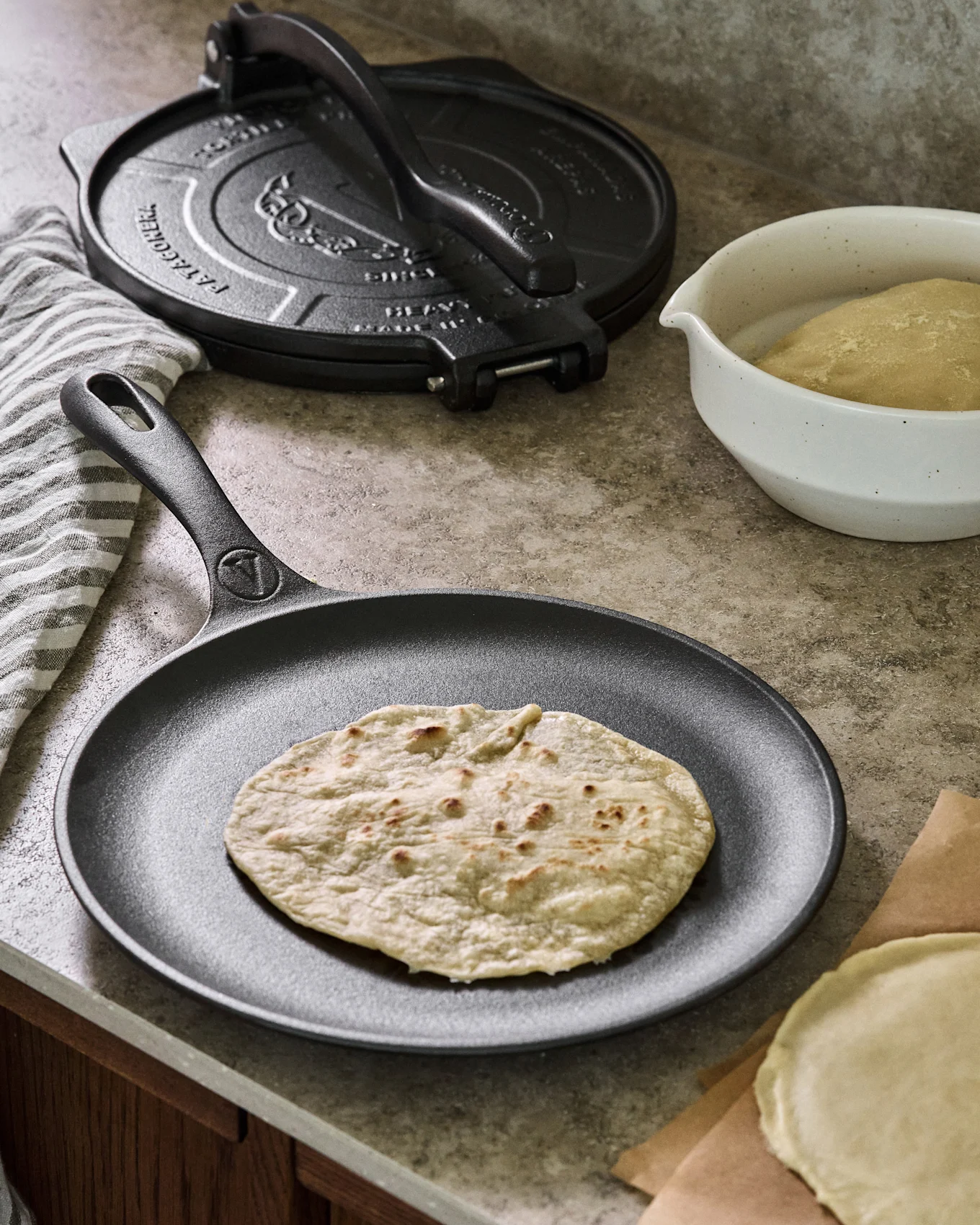 Eine gekochte Tortilla liegt auf einer schwarzen Gusseisenpfanne, mit einer Tortillapresse, Teigschüssel und ungekochten Tortillas im Hintergrund.