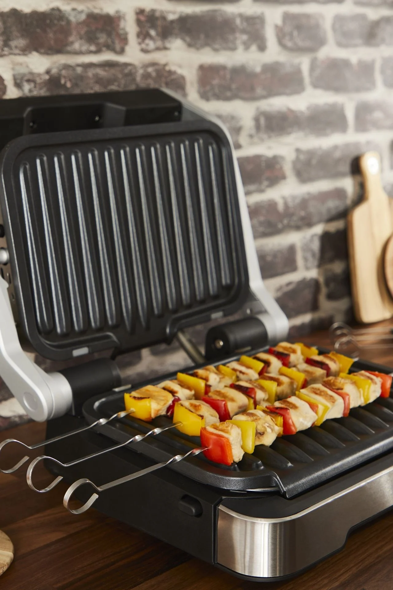 Ein geöffneter Elektrogrill brät Spieße mit Hähnchen und roten sowie gelben Paprika auf einer Holztheke.