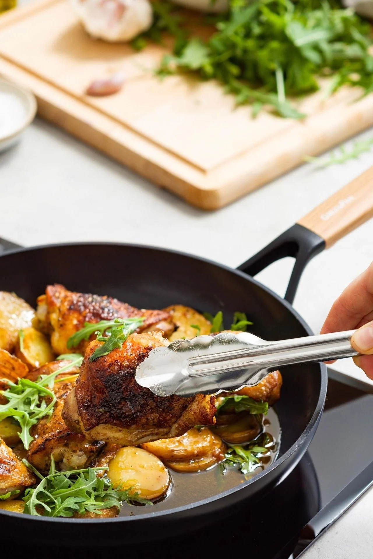 Hand mit Zange wendet knuspriges Hähnchenschenkel in schwarzer Pfanne mit Bratkartoffeln und Rucola.