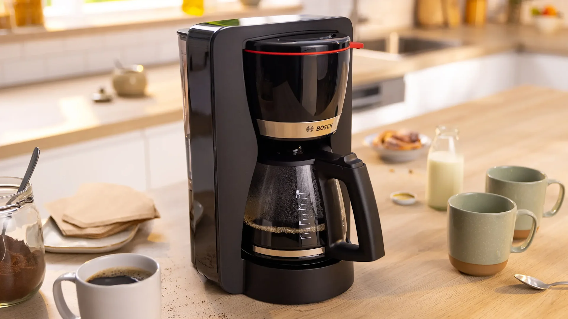 Eine schwarze Filterkaffeemaschine auf einer Holztheke mit Kaffee, Tassen und Milch, die eine Frühstücksszene andeutet.