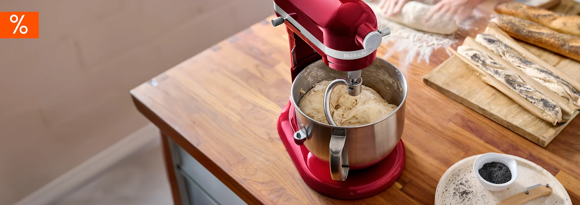Roter Standmixer knetet Teig auf einem Holztisch. Baguettes, Mohn und ein Angebotssymbol sind sichtbar.