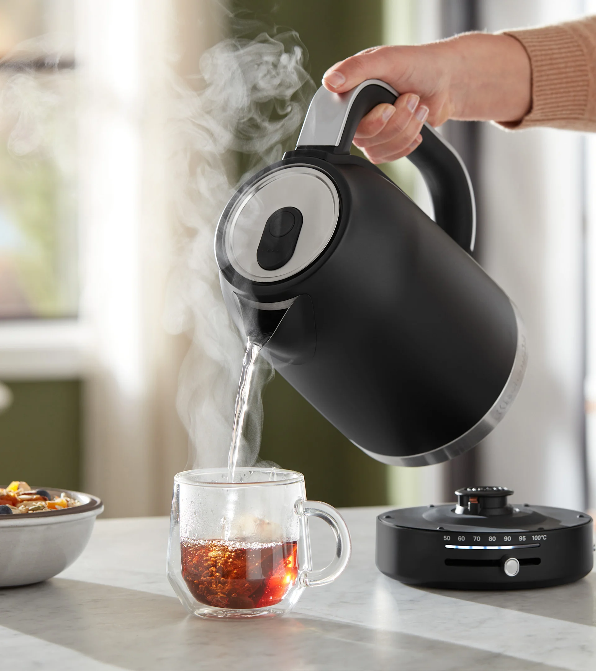 Hand gießt heißes Wasser aus einem schwarzen Wasserkocher mit Temperaturregelung in eine dampfende Teetasse.