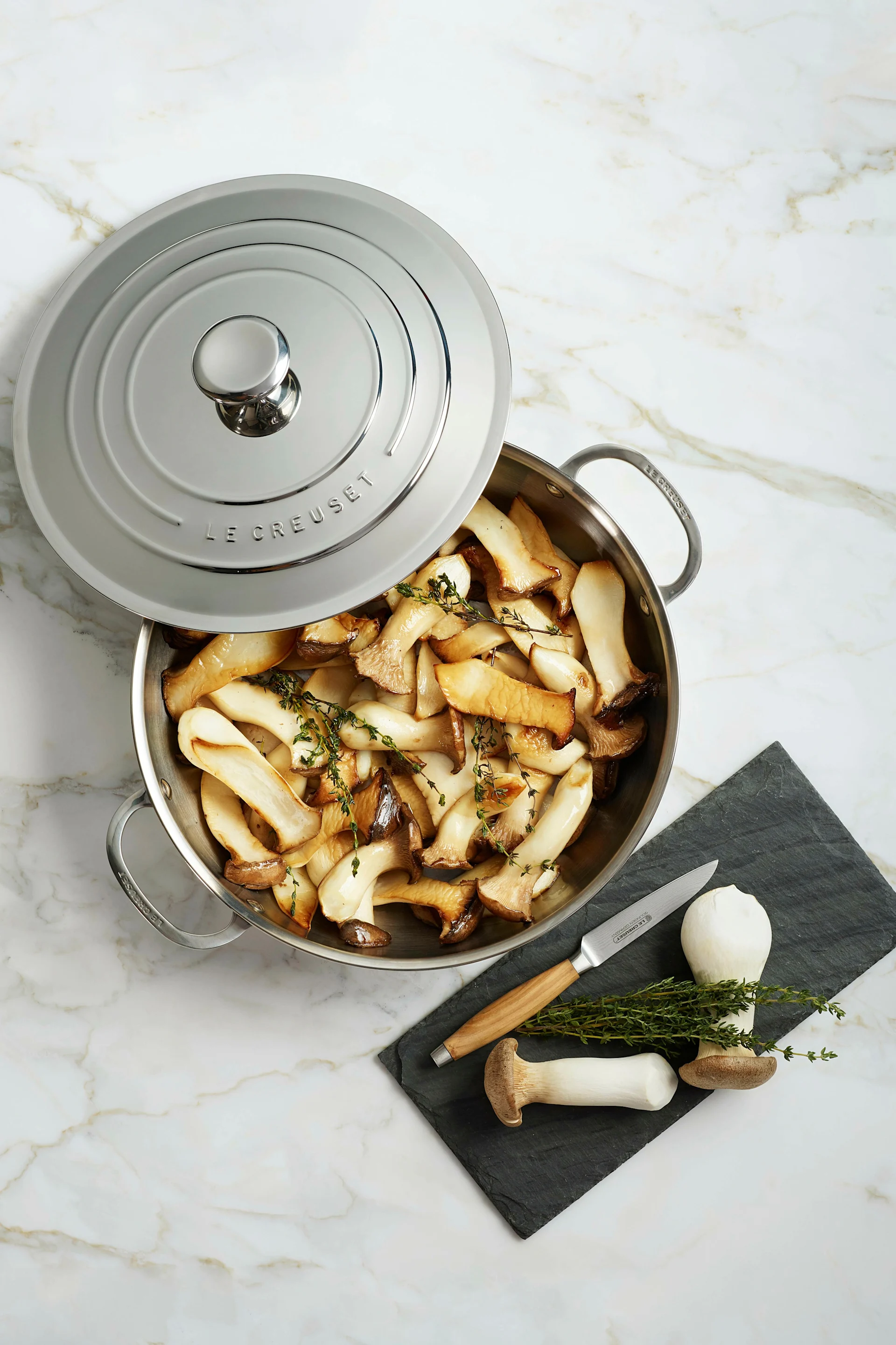 Ein Edelstahltopf mit gebratenen Kräuterseitlingen und Thymian, teilweise vom Deckel bedeckt. Daneben liegen rohe Pilze, Knoblauch und ein Messer auf einem Schieferbrett.