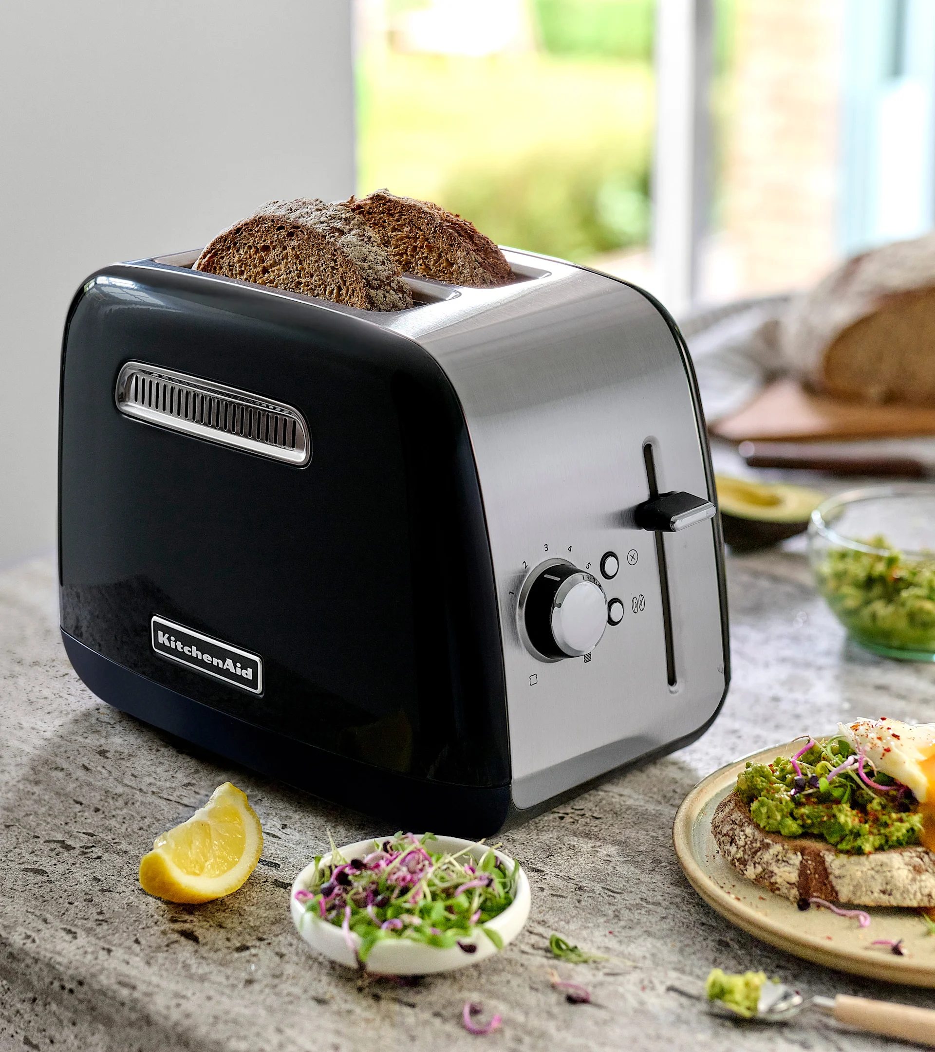 Ein schwarzer und edelstahlfarbener KitchenAid Toaster mit zwei Scheiben dunklem Brot. Auf der Theke Avocadotoast mit Ei, Sprossen und einer Zitronenspalte.